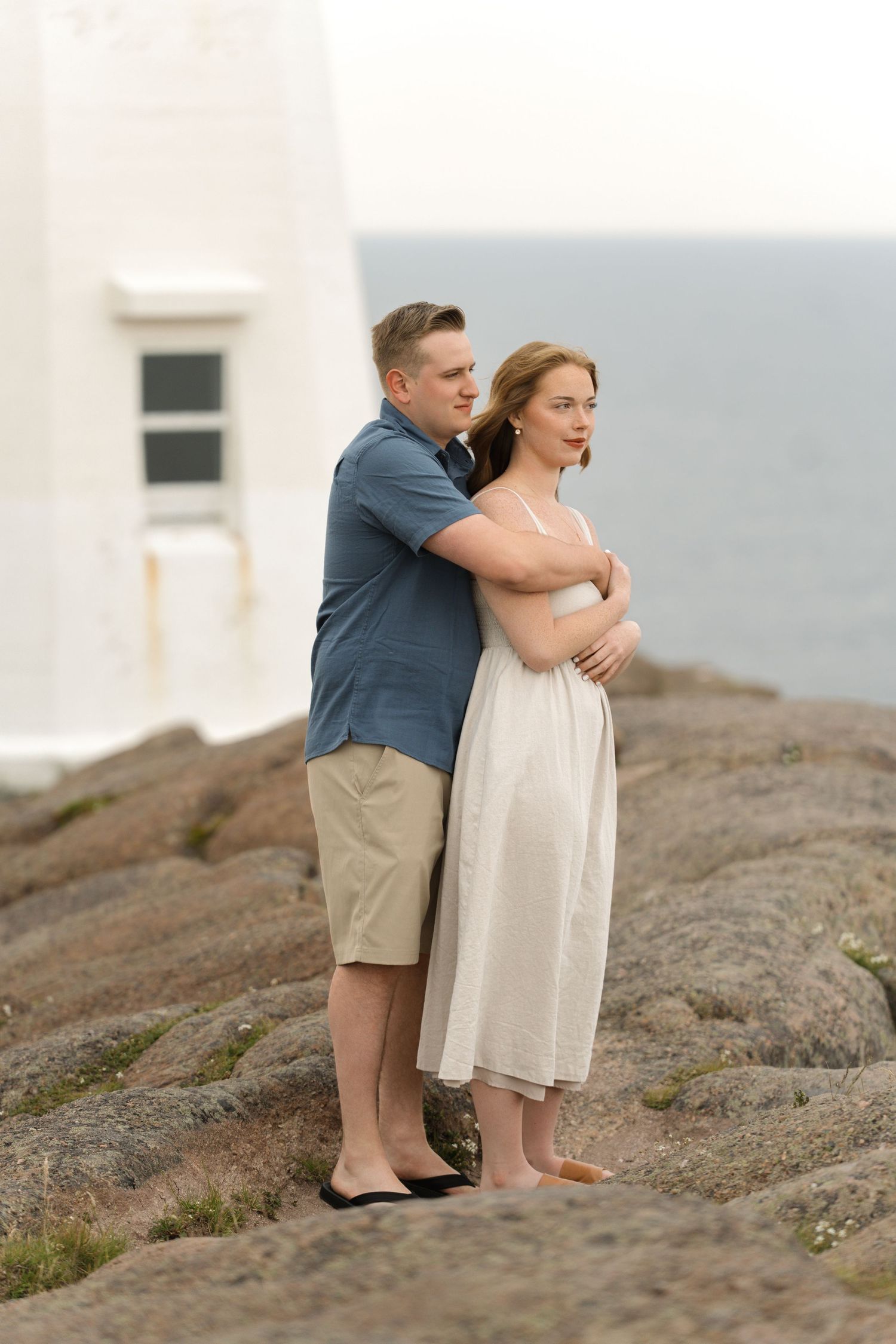 Romantic couple embraces while standing together on rocky coastal terrain near a white lighthouse.