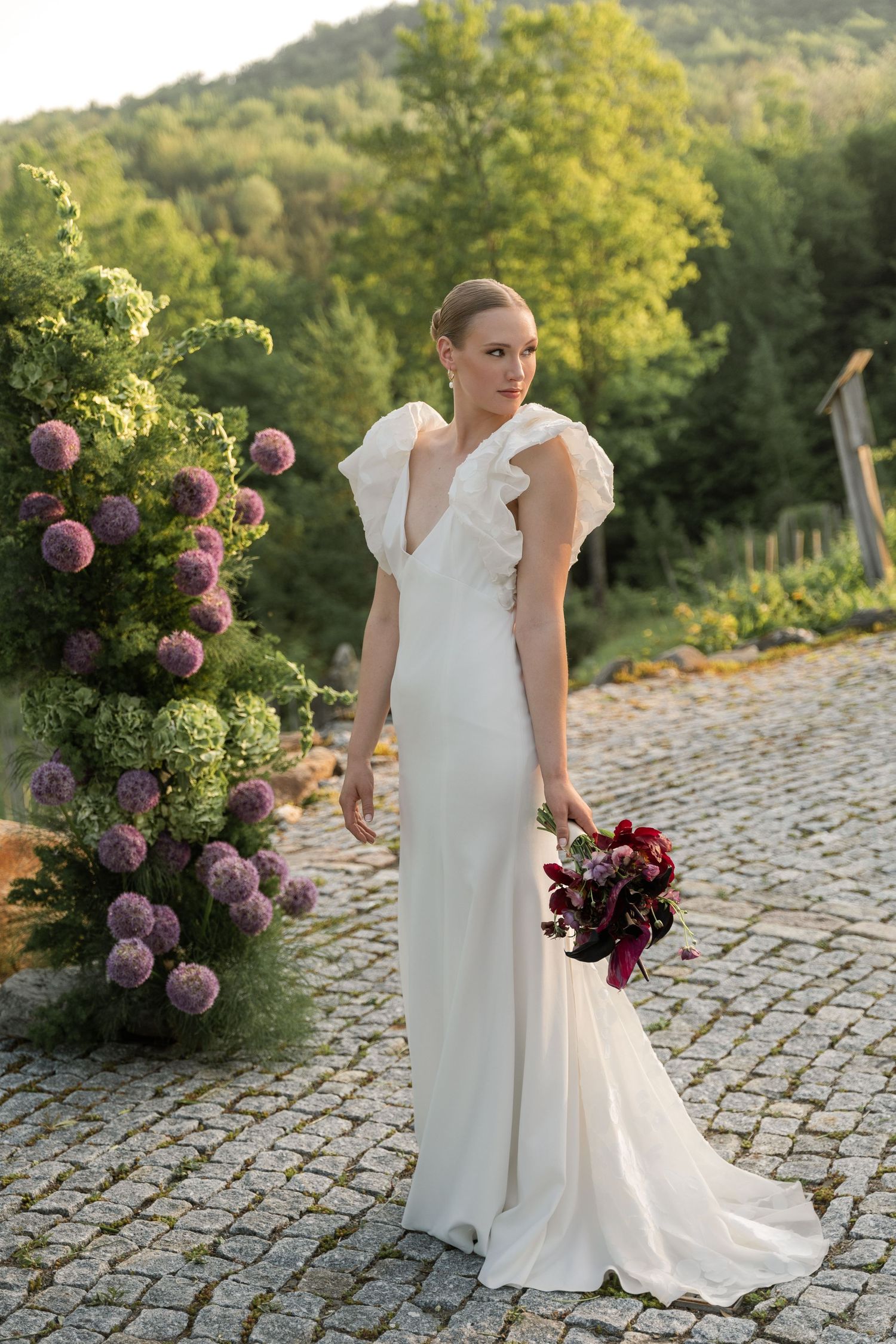 A bride in a white wedding dress stands on stone pavers near blooming purple allium flowers in a garden setting.