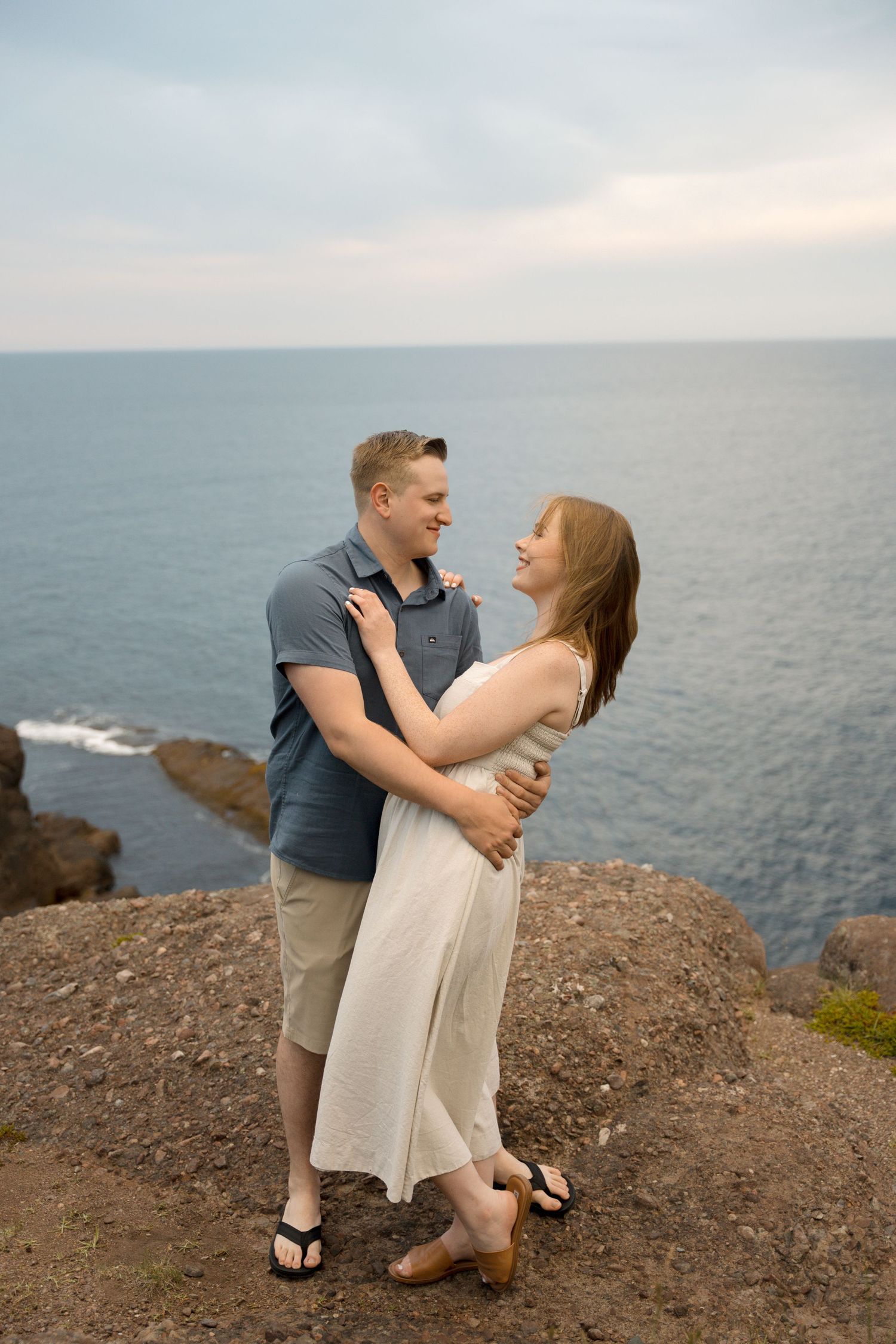 A couple shares a joyful embrace on a coastal cliff overlooking the ocean during sunset.