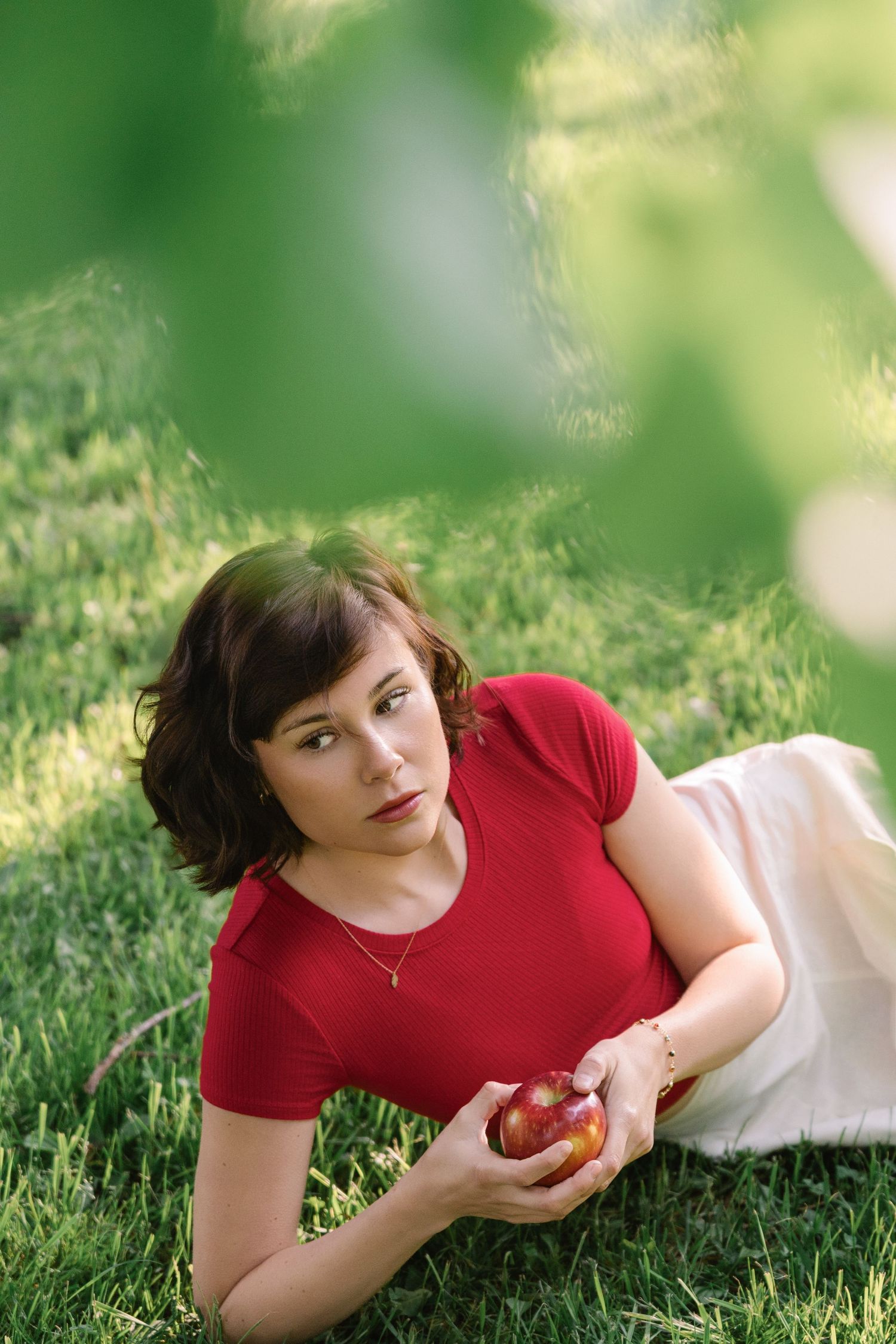 A person in a red shirt lies in green grass holding a red apple on a sunny summer day.