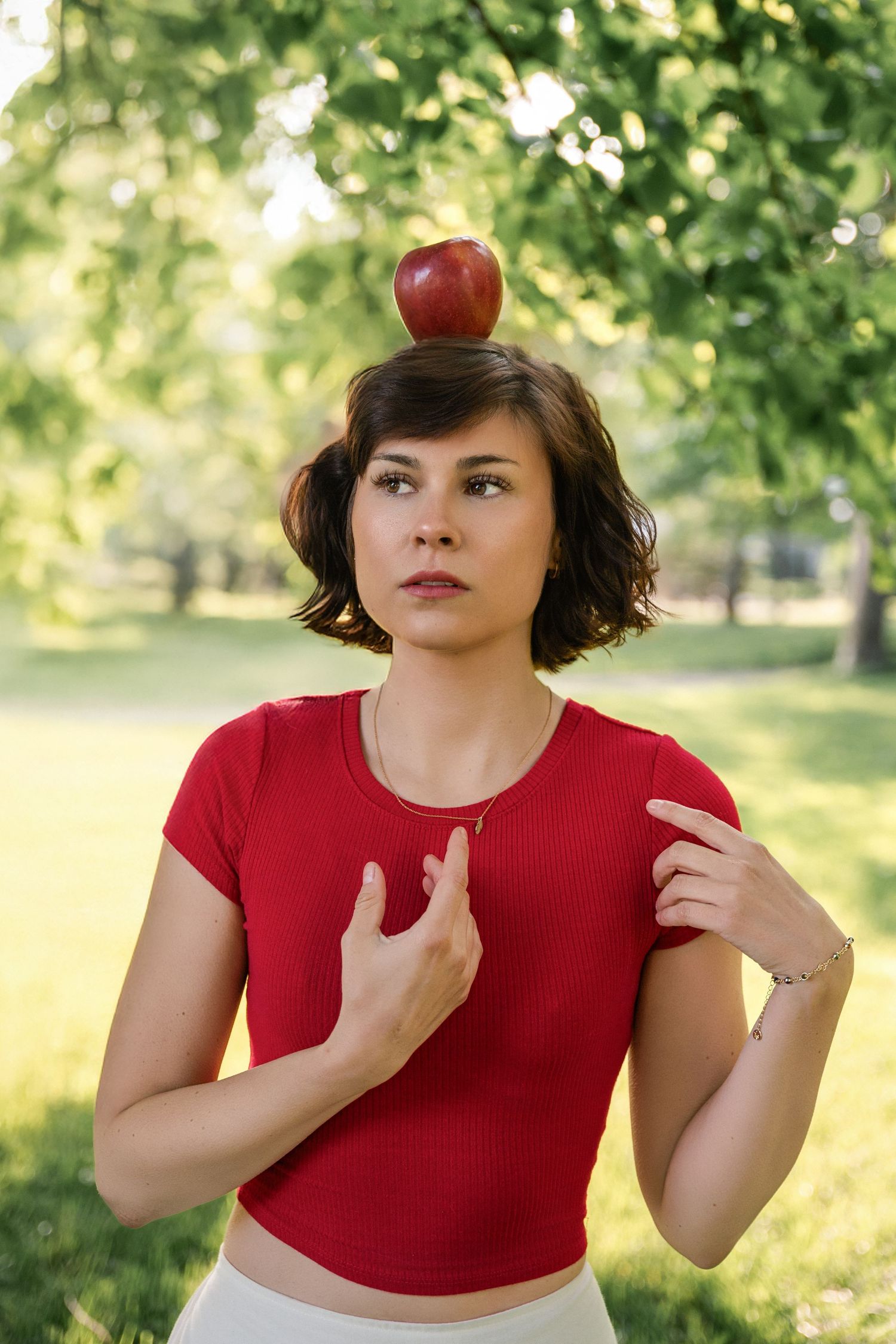 A red apple balances on a person's head while they pose in a red shirt outdoors with trees in the background.