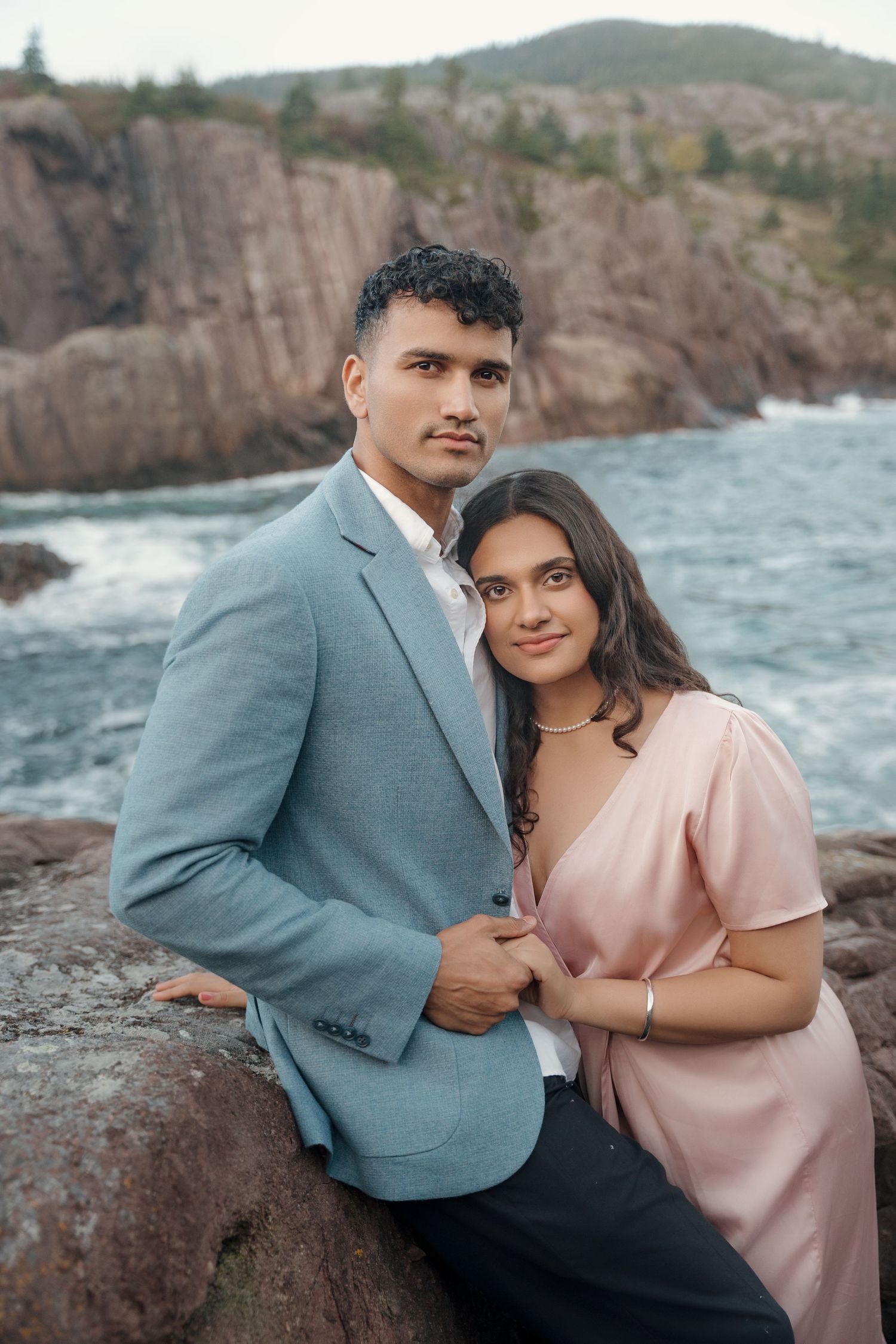 A couple in formal attire poses together on rocky cliffs overlooking the ocean at sunset.