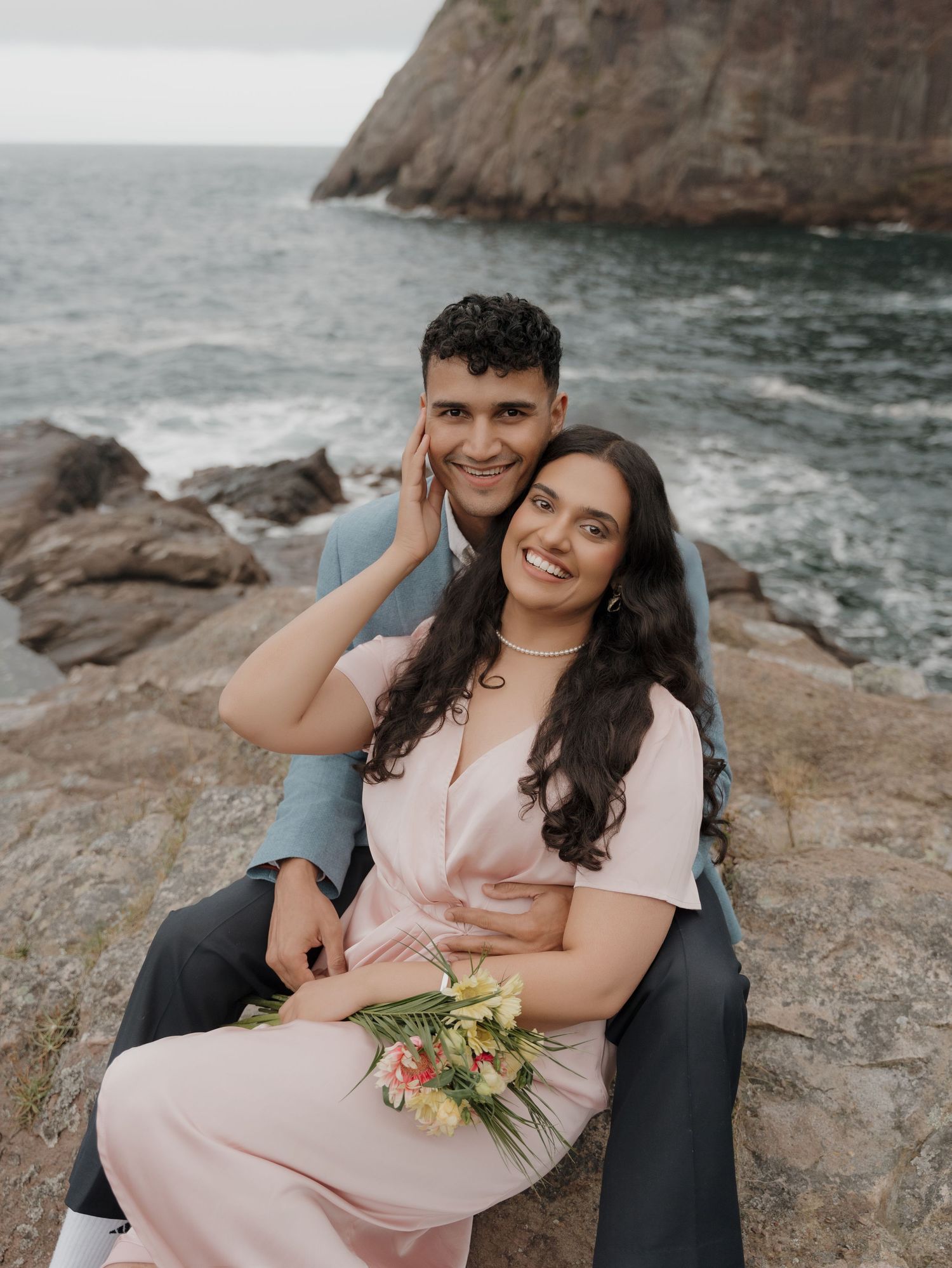 A couple enjoys a romantic moment together on rocky coastal cliffs overlooking the ocean at sunset.