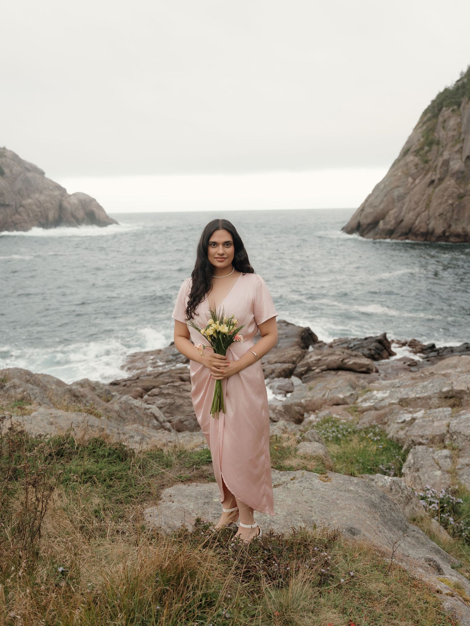 Pink dress flows in the ocean breeze against rocky coastal cliffs and stormy seas.