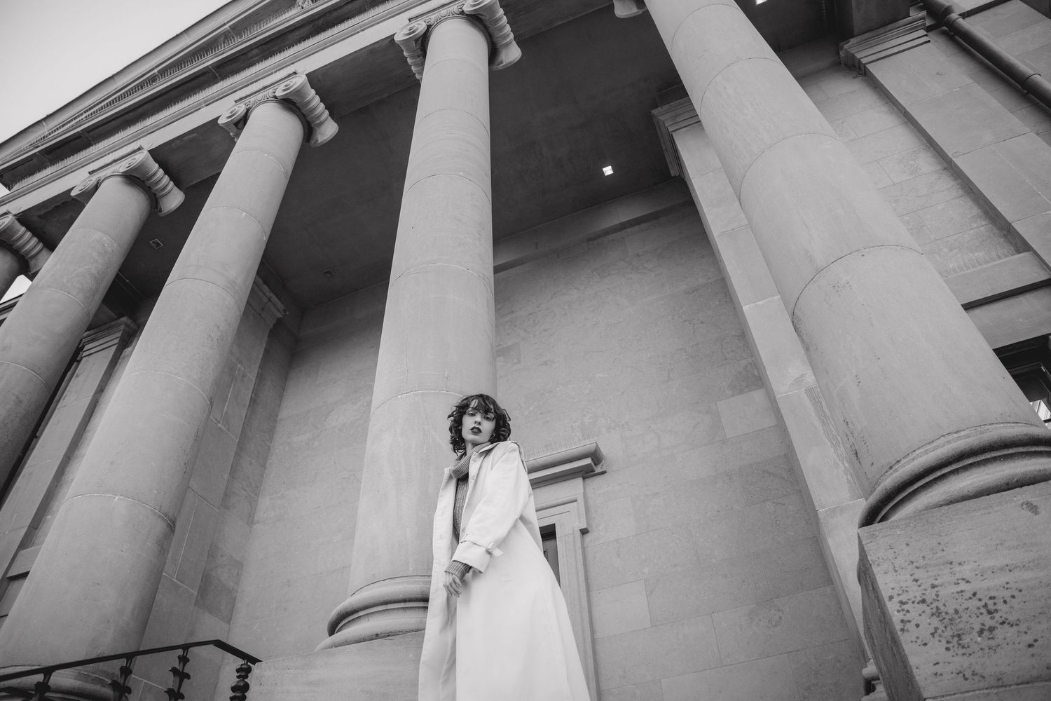 Low angle black and white shot of person in dress standing among massive classical columns.