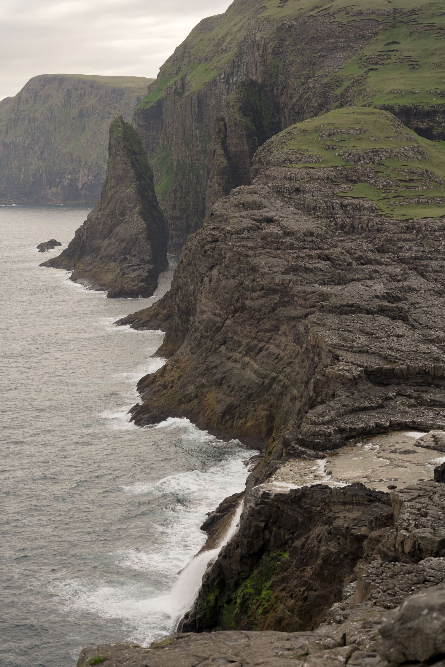 Dramatic rocky sea cliffs with green tops meet crashing waves along a rugged coastline on an overcast day.