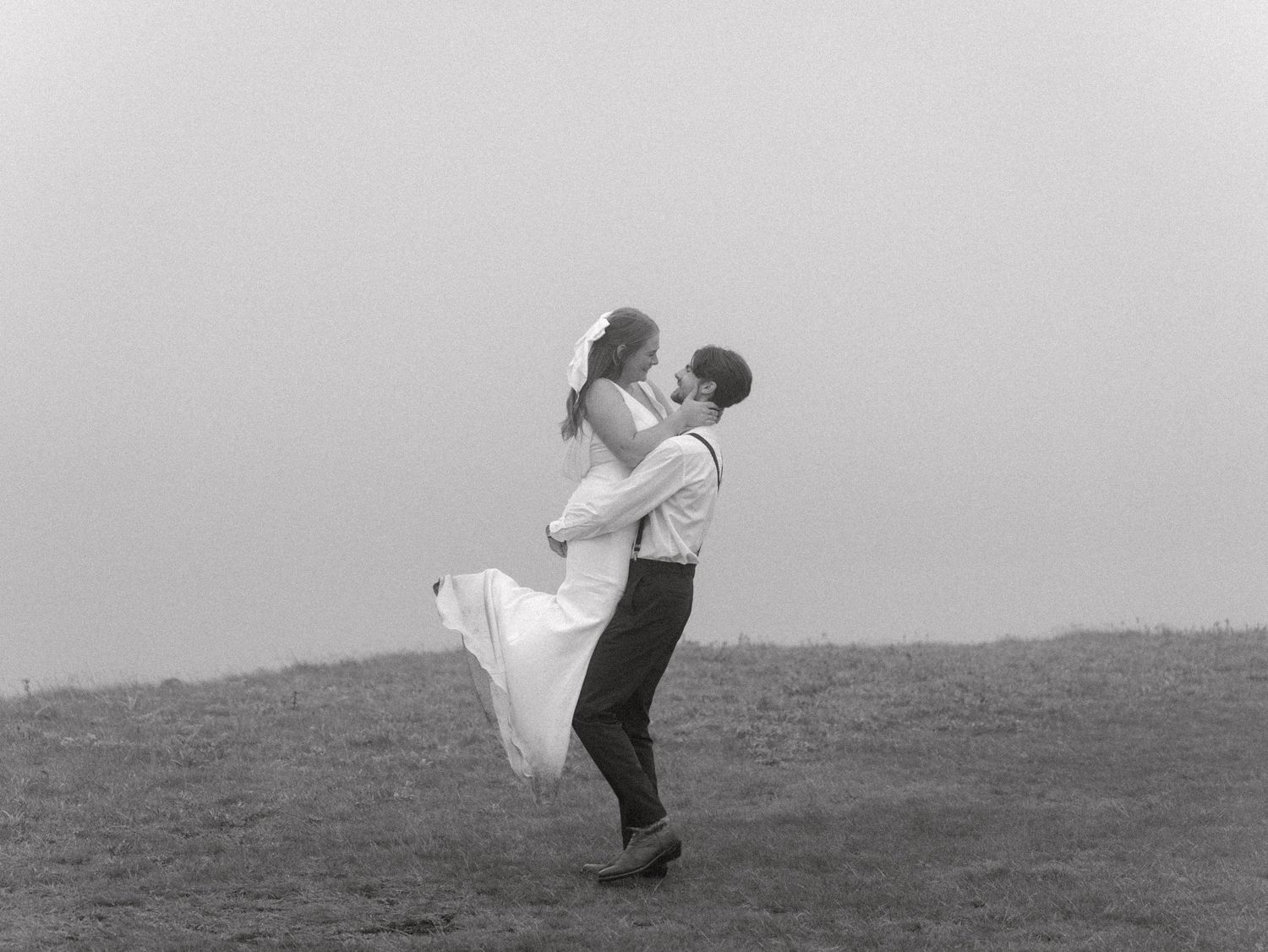A couple in wedding attire dances romantically on a hillside in black and white photography.