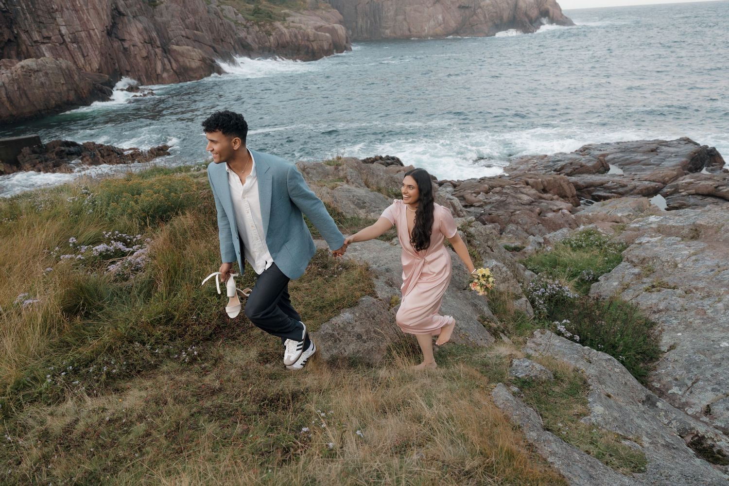 Couple exploring rocky coastal cliffs overlooking the ocean during sunset while capturing scenic photos.