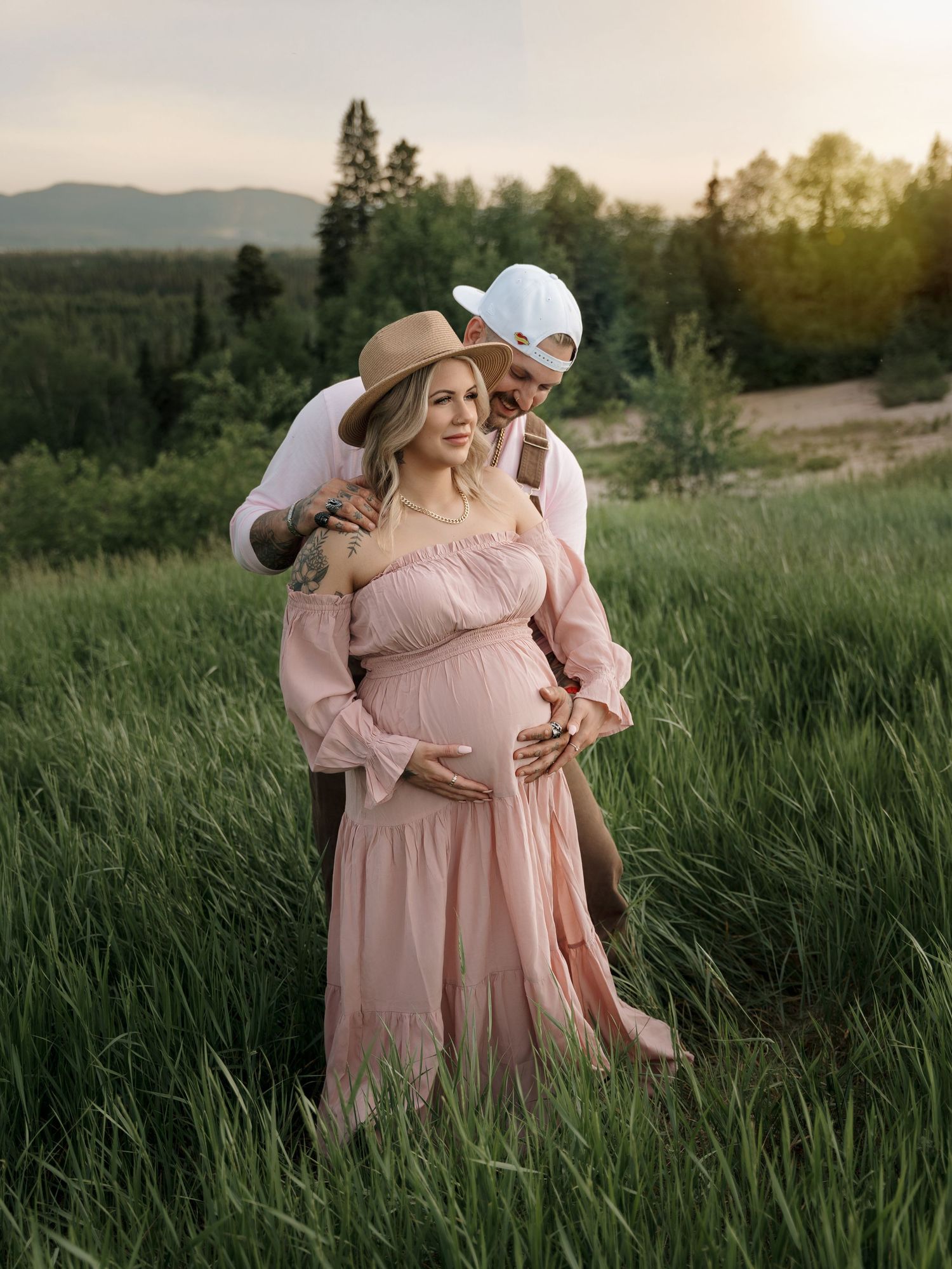 Couple in pink dress and white hat embracing in tall grass at sunset.