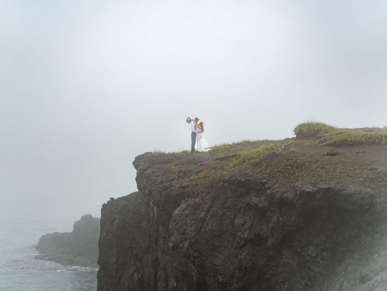 Two figures stand on a misty cliff edge overlooking the rocky coastline during a foggy day.