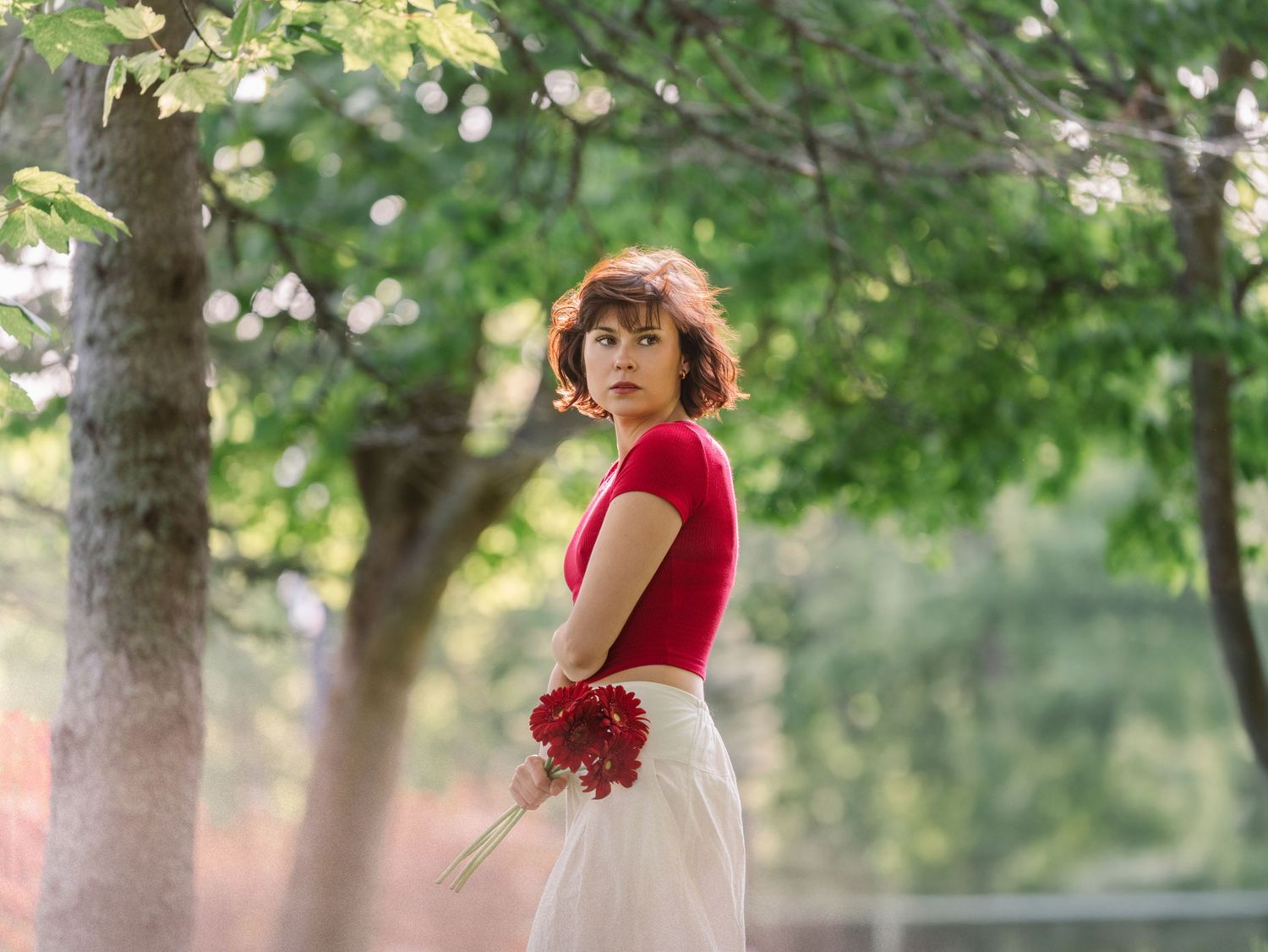 A person in a red dress poses elegantly in a lush green forest setting during a photo shoot.