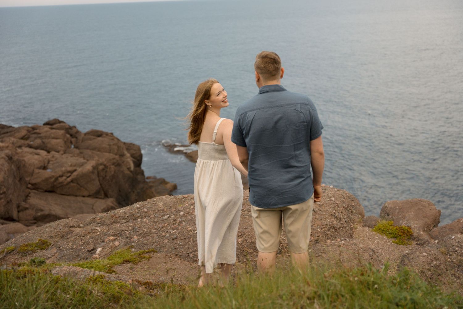 A couple stands together on a rocky coastal cliff overlooking a calm ocean on an overcast day.