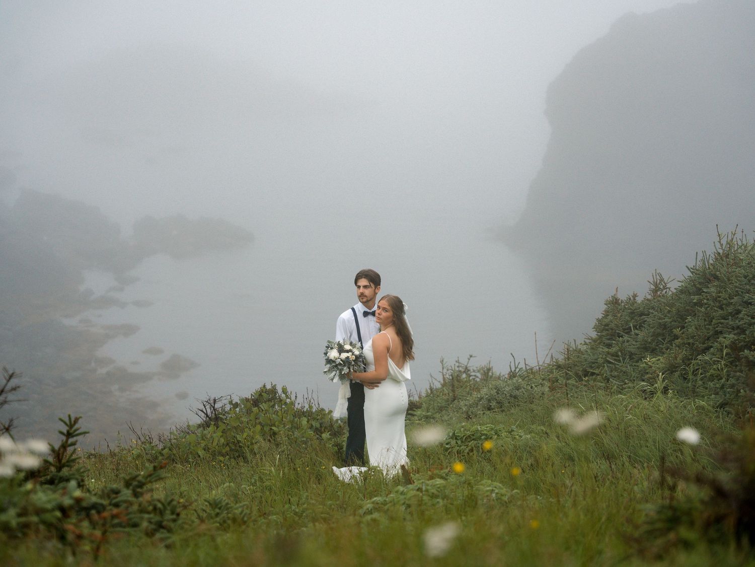A wedding couple stands on a misty mountaintop meadow surrounded by wildflowers and evergreen trees.