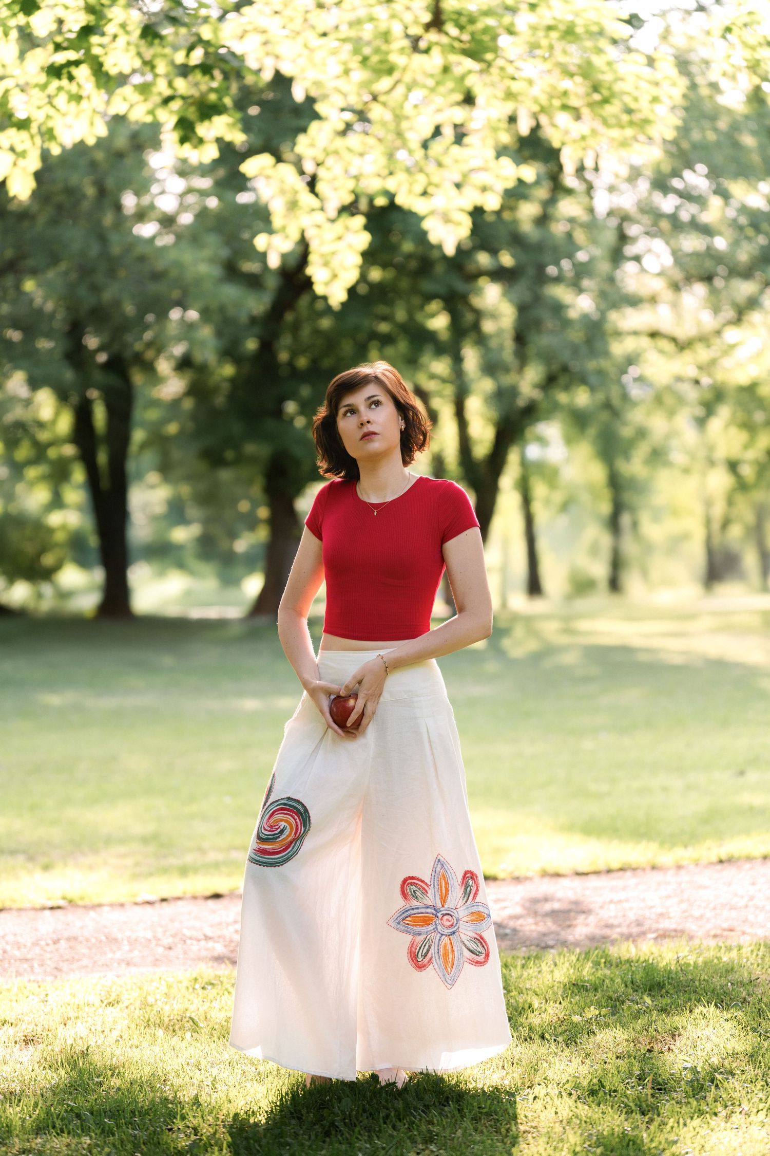 Woman in red top and white skirt with butterfly detail twirls gracefully in a sunlit park setting.