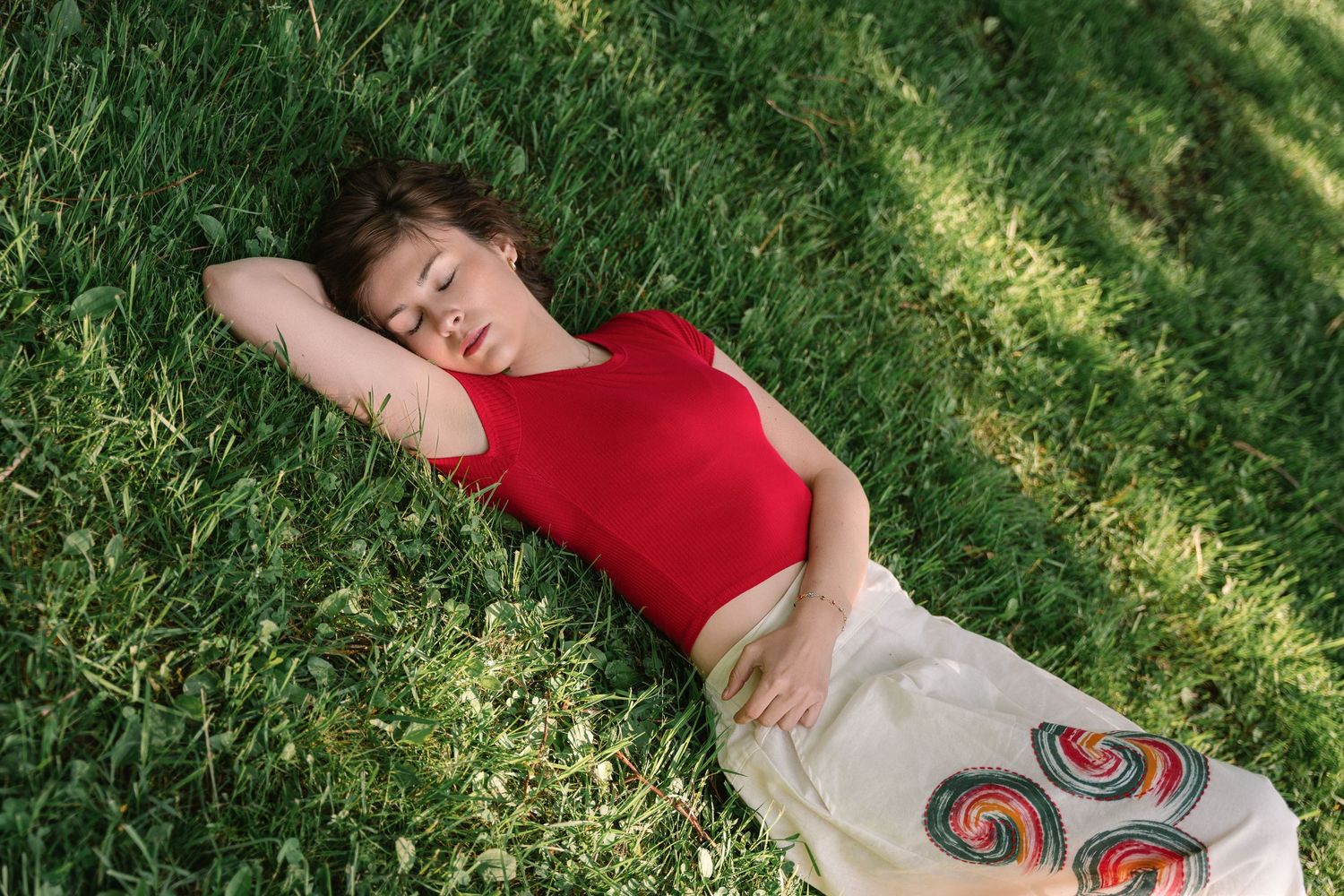 A person in a red tank top lies peacefully on green grass in the sunlight.