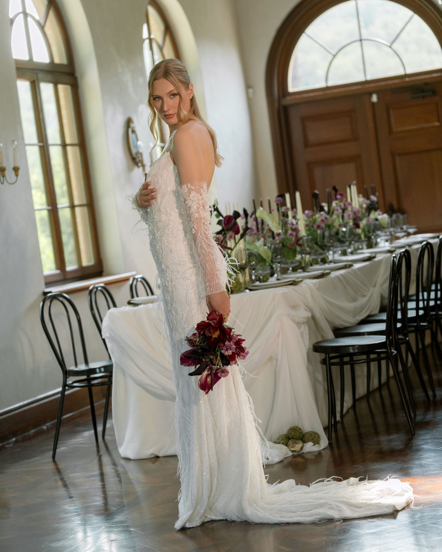 Bride in elegant lace wedding gown poses gracefully in a grand ballroom with arched windows and reception tables.