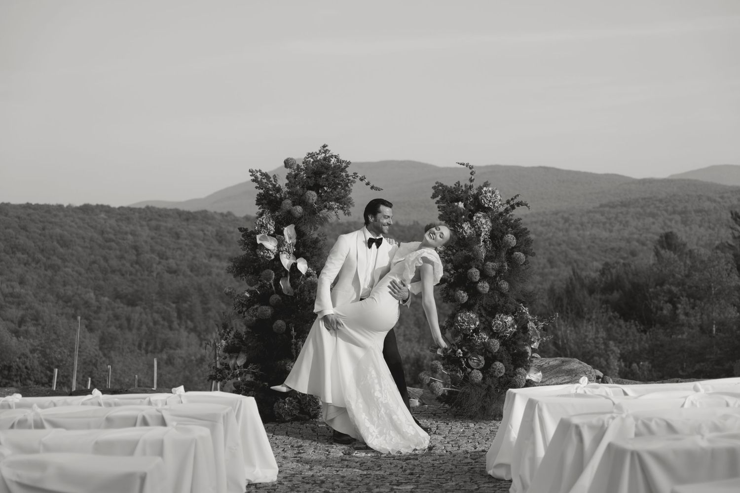 A couple dances joyfully at an outdoor mountain wedding ceremony with a floral arch and chairs set up for guests.