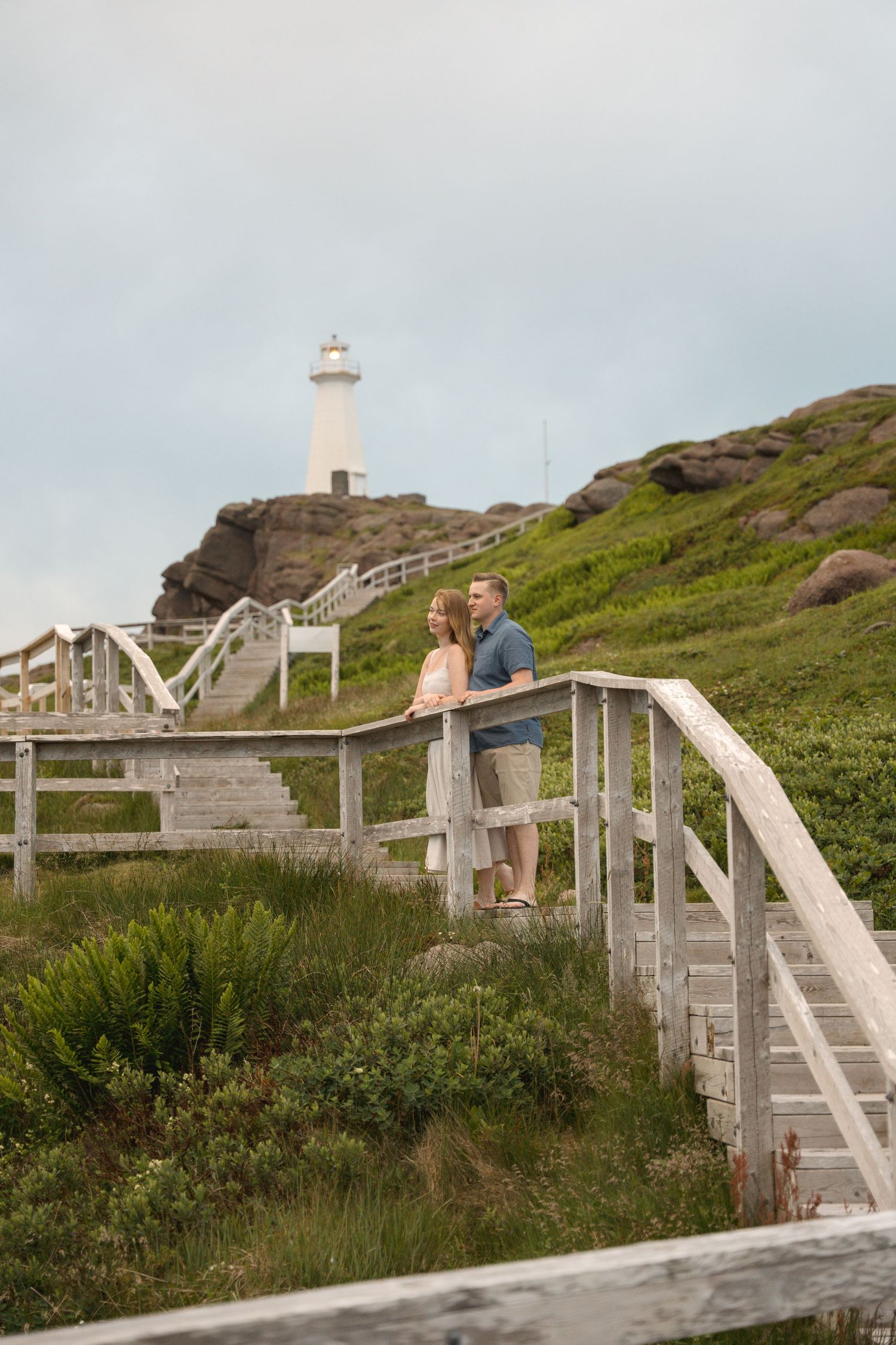 A wooden stairway leads up a green coastal hillside to a lighthouse on the cliff's edge.