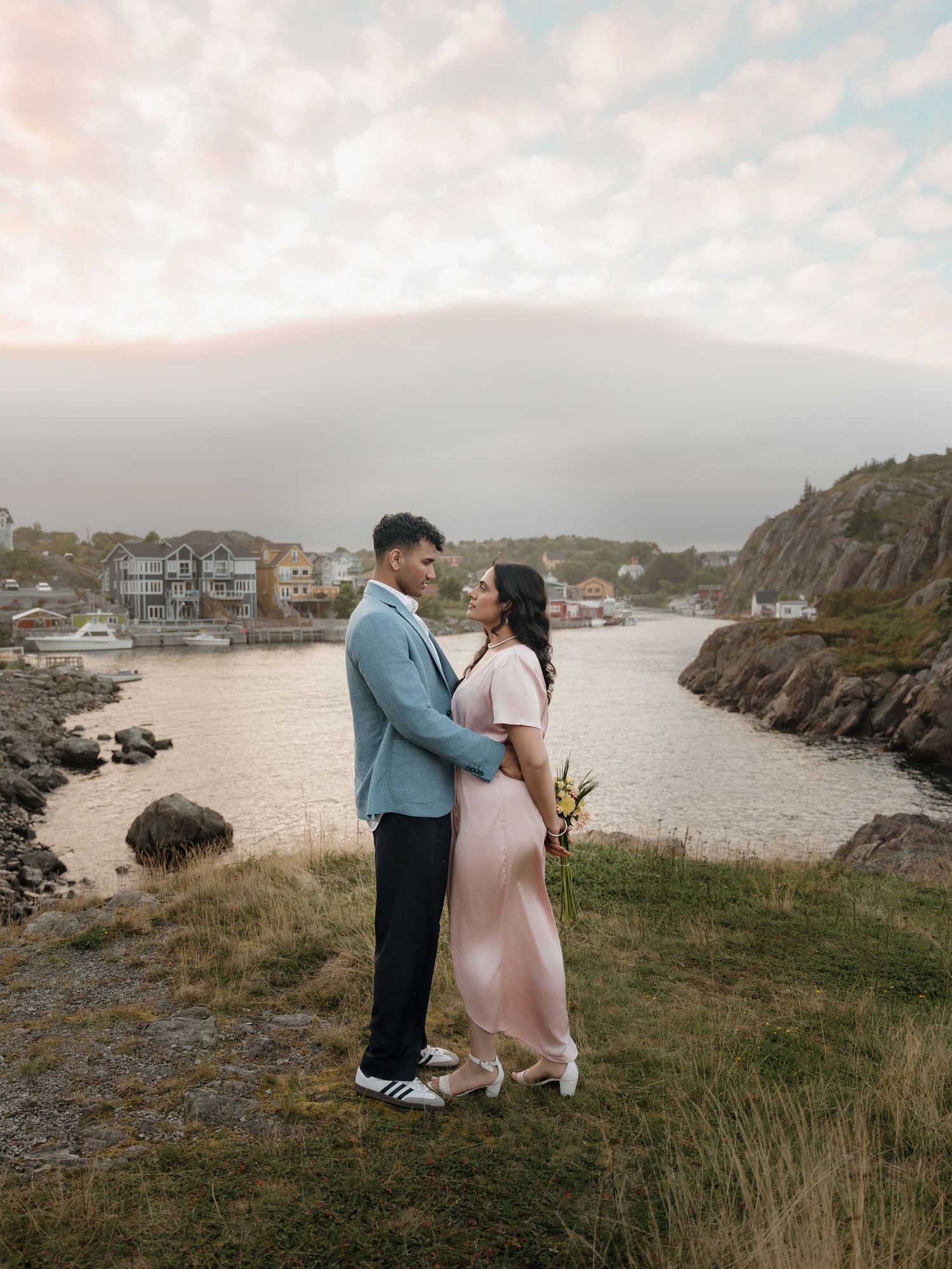 A couple embraces on a rocky coastal cliff overlooking a serene harbor at sunset.