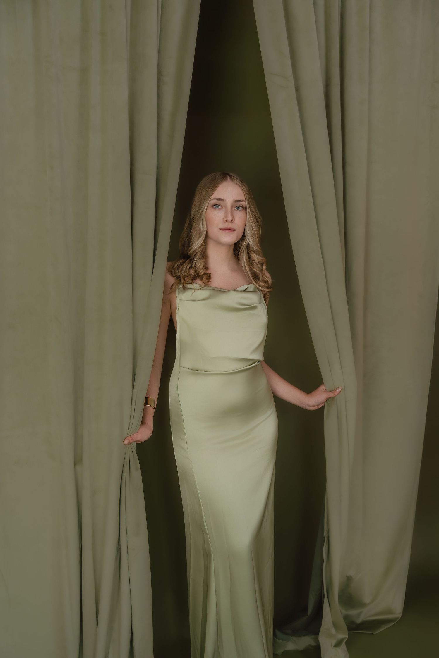 Elegant satin dress in light sage green photographed against matching curtains in moody studio lighting.