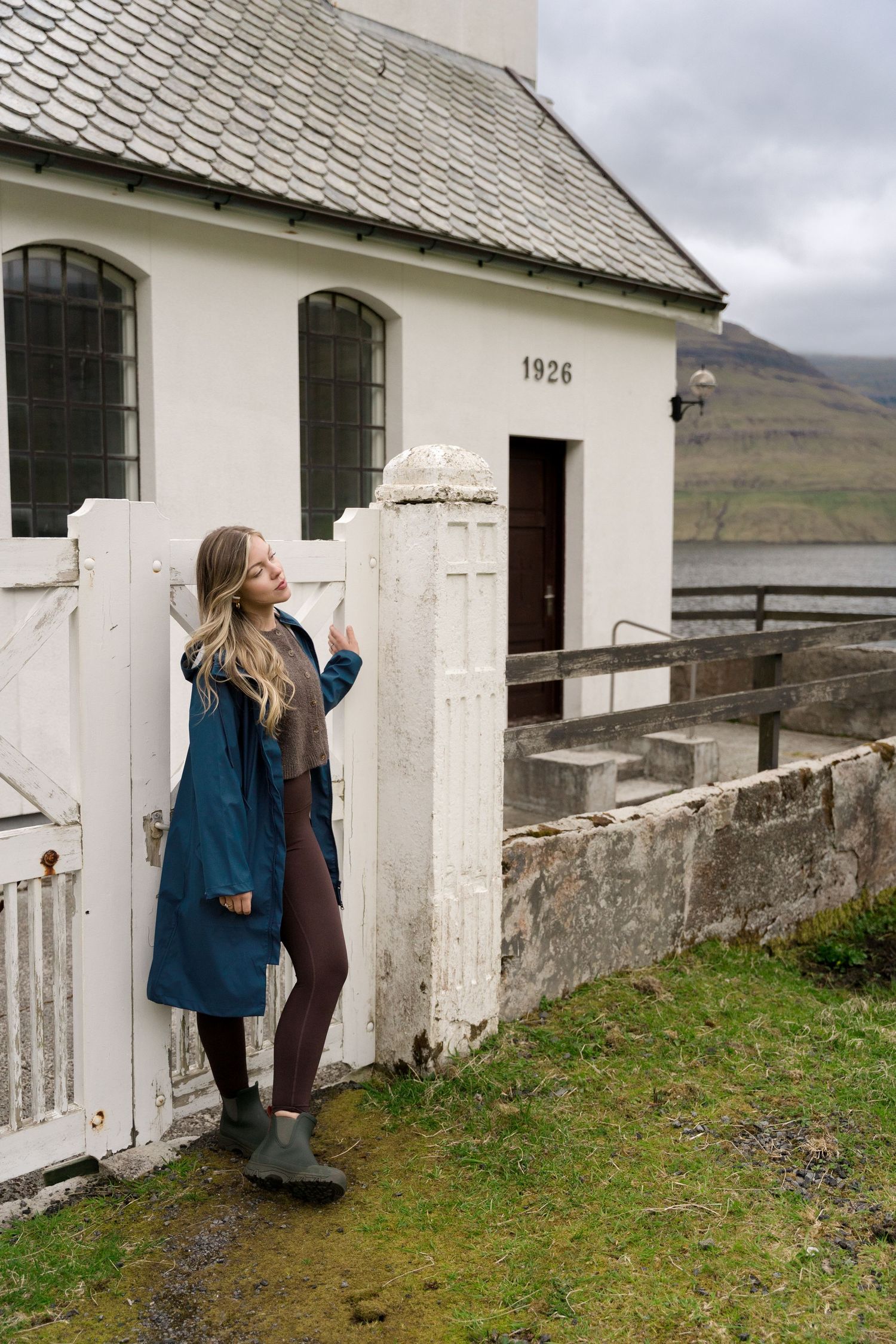 Woman in teal coat leaning against white gate of historic 1926 church with fjord views in background.