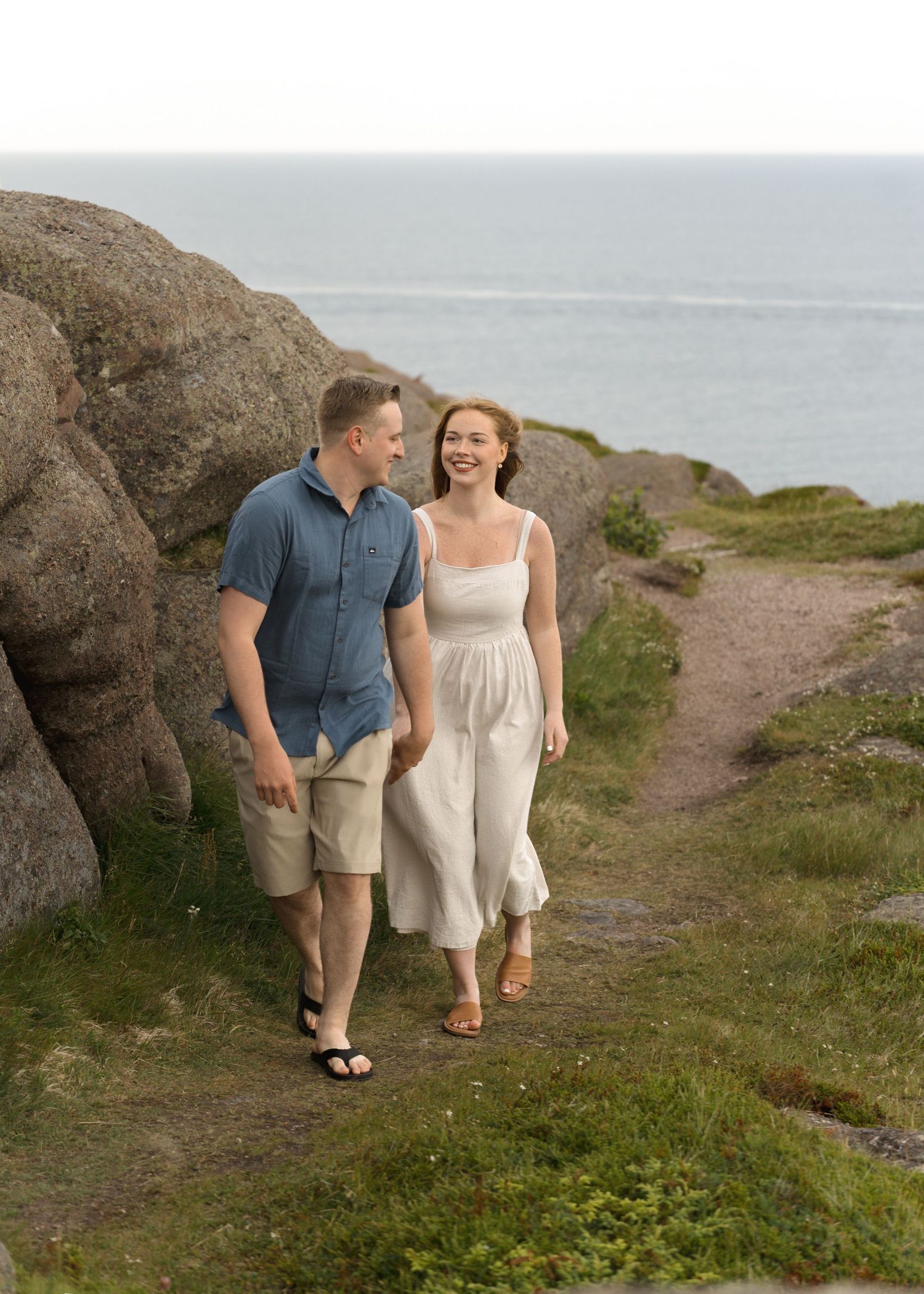 A couple walks together on a coastal path with large rocks and ocean views in the background.