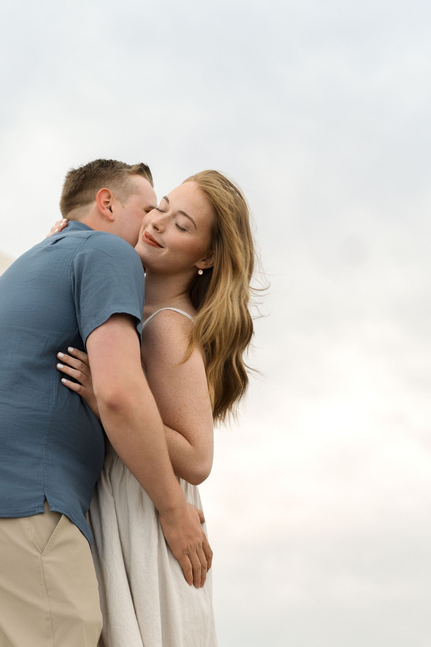 A romantic couple sharing tender kisses while embracing against a light cloudy sky background.