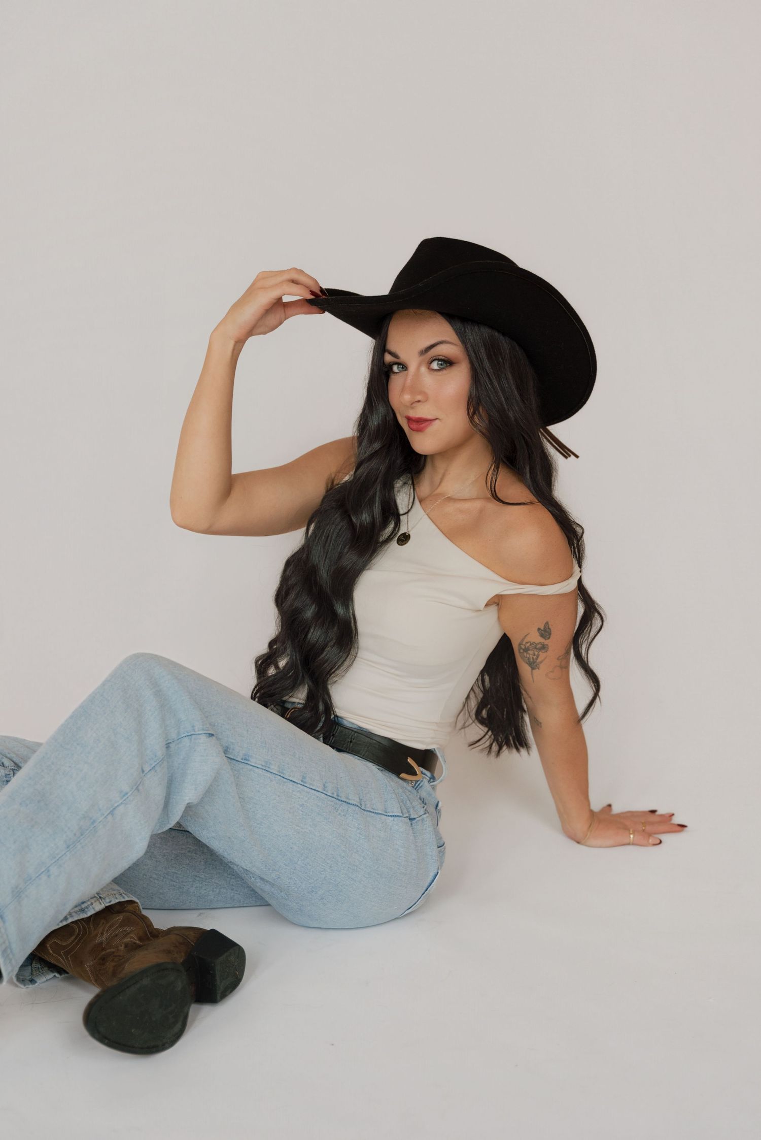 Model in white tank top, light jeans and black hat poses on white background in studio photoshoot.