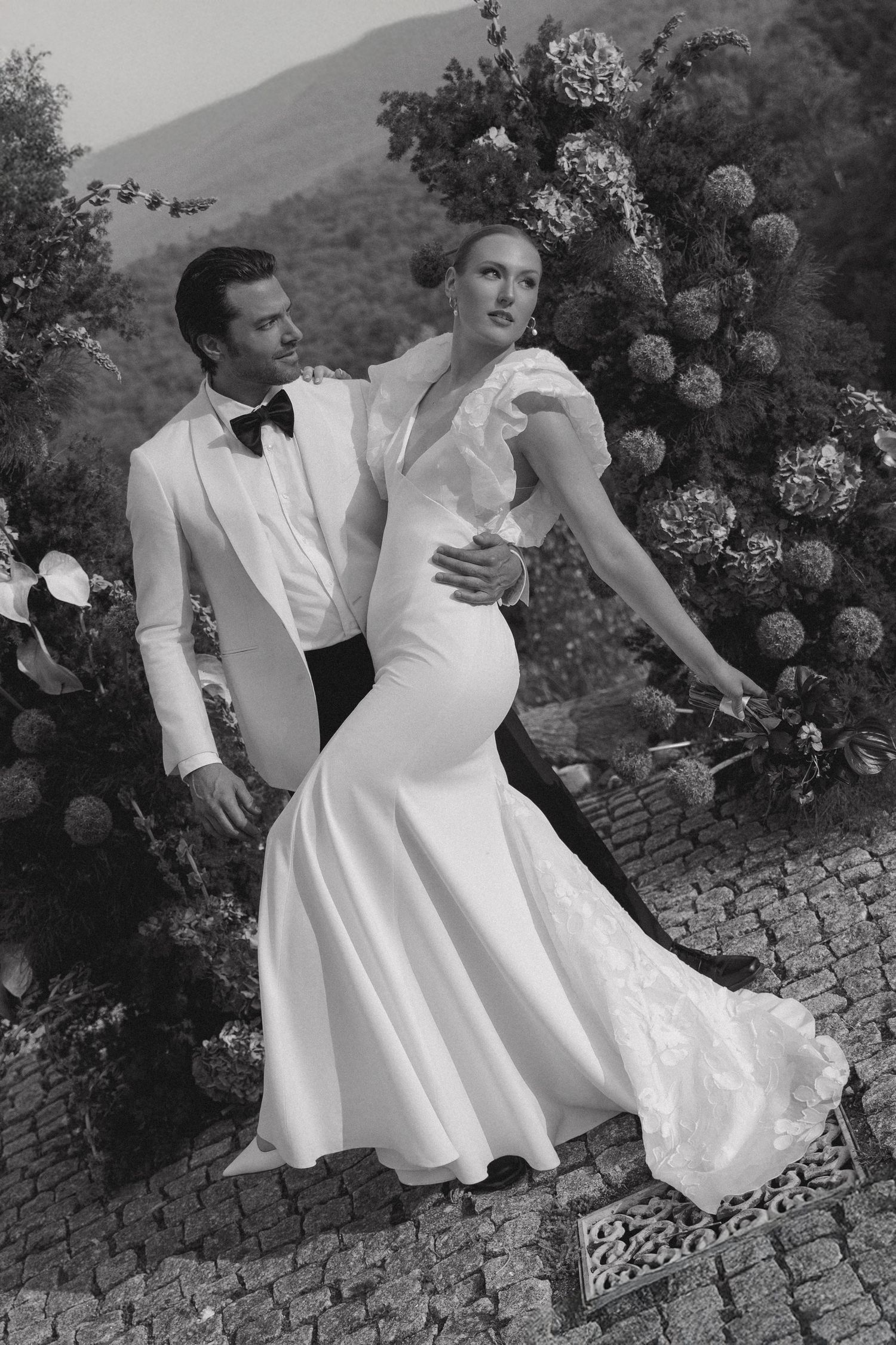A black and white photograph of a romantic couple in formal attire posing outdoors on a stone path with blooming flowers.