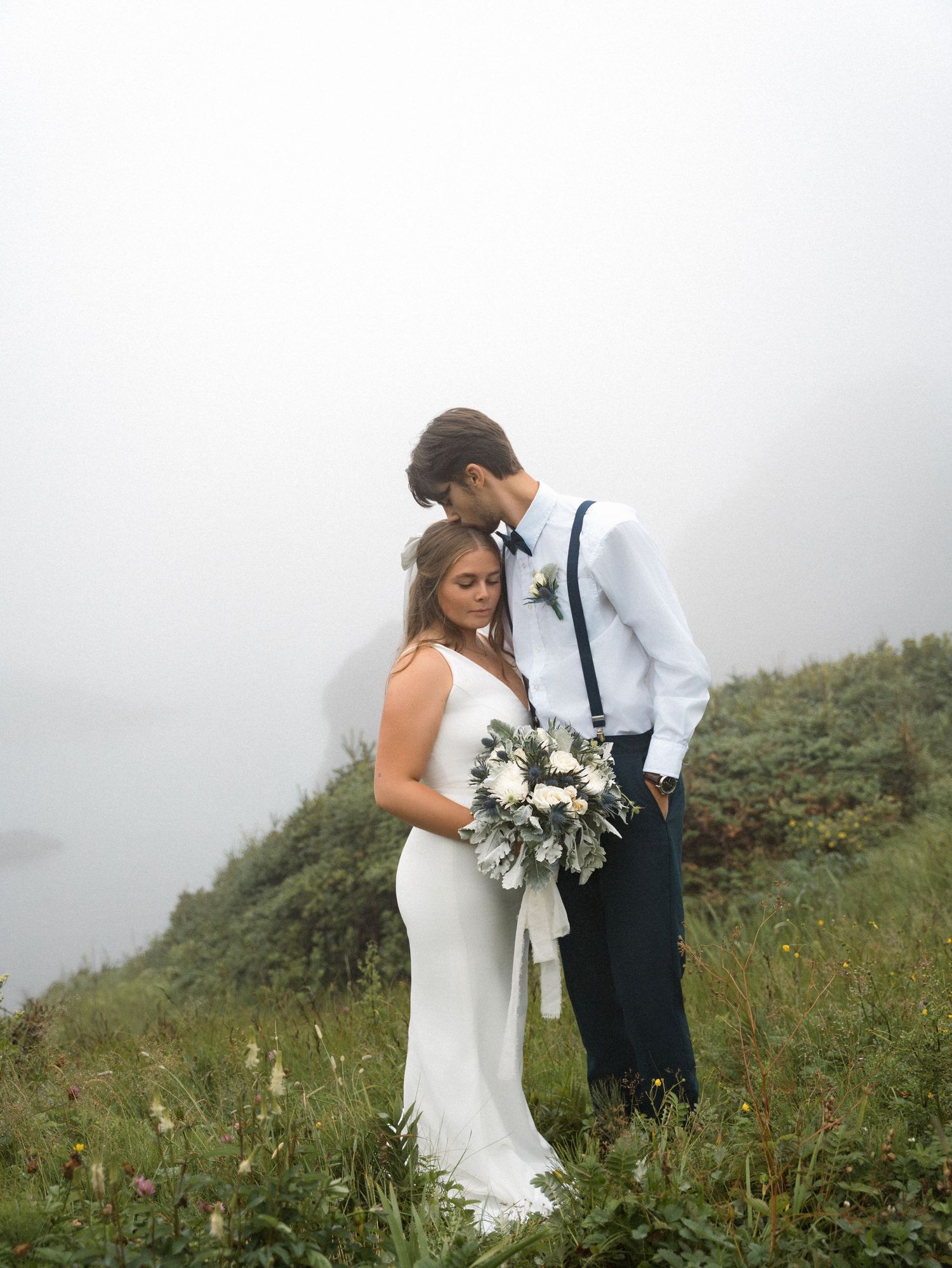 A couple shares an intimate moment on a misty hilltop in wedding attire with a white bouquet.