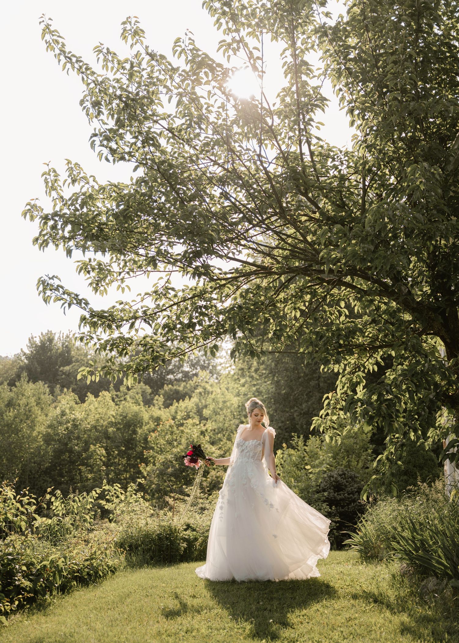 A white wedding dress twirls gracefully under a sunlit tree in a lush garden setting.