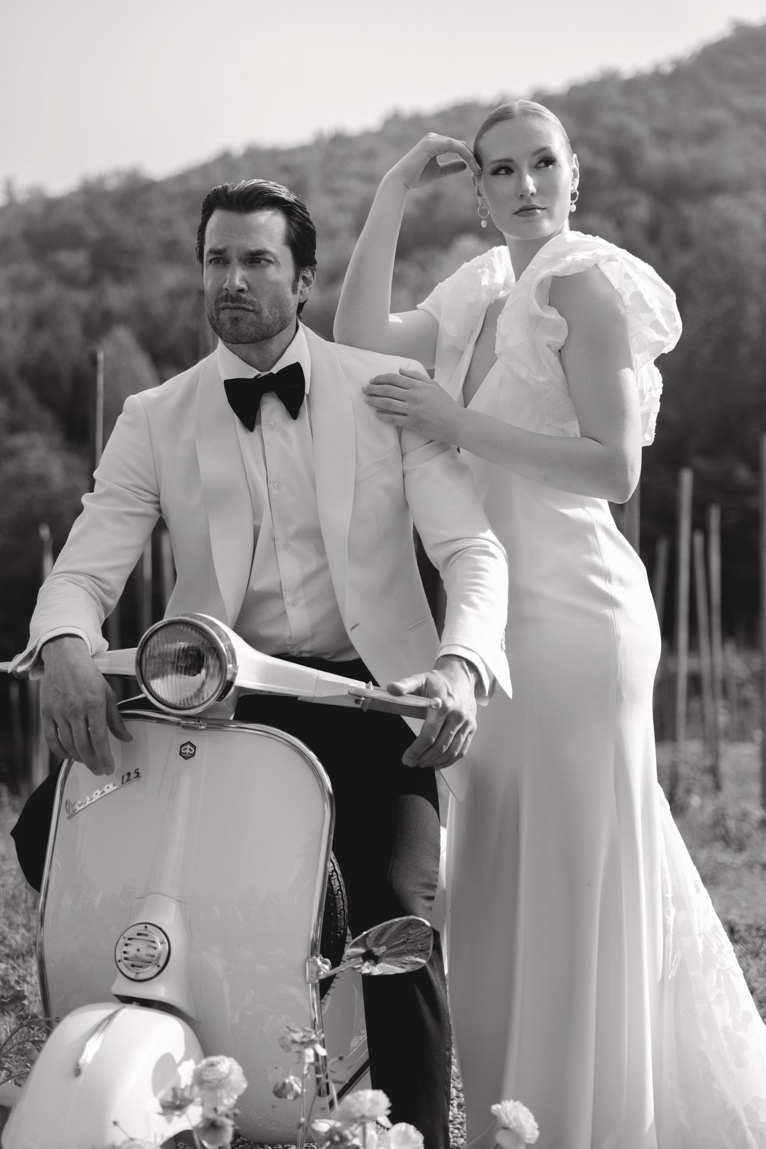 Black and white fashion photo featuring a vintage Vespa scooter with couple in elegant formalwear posing outdoors.