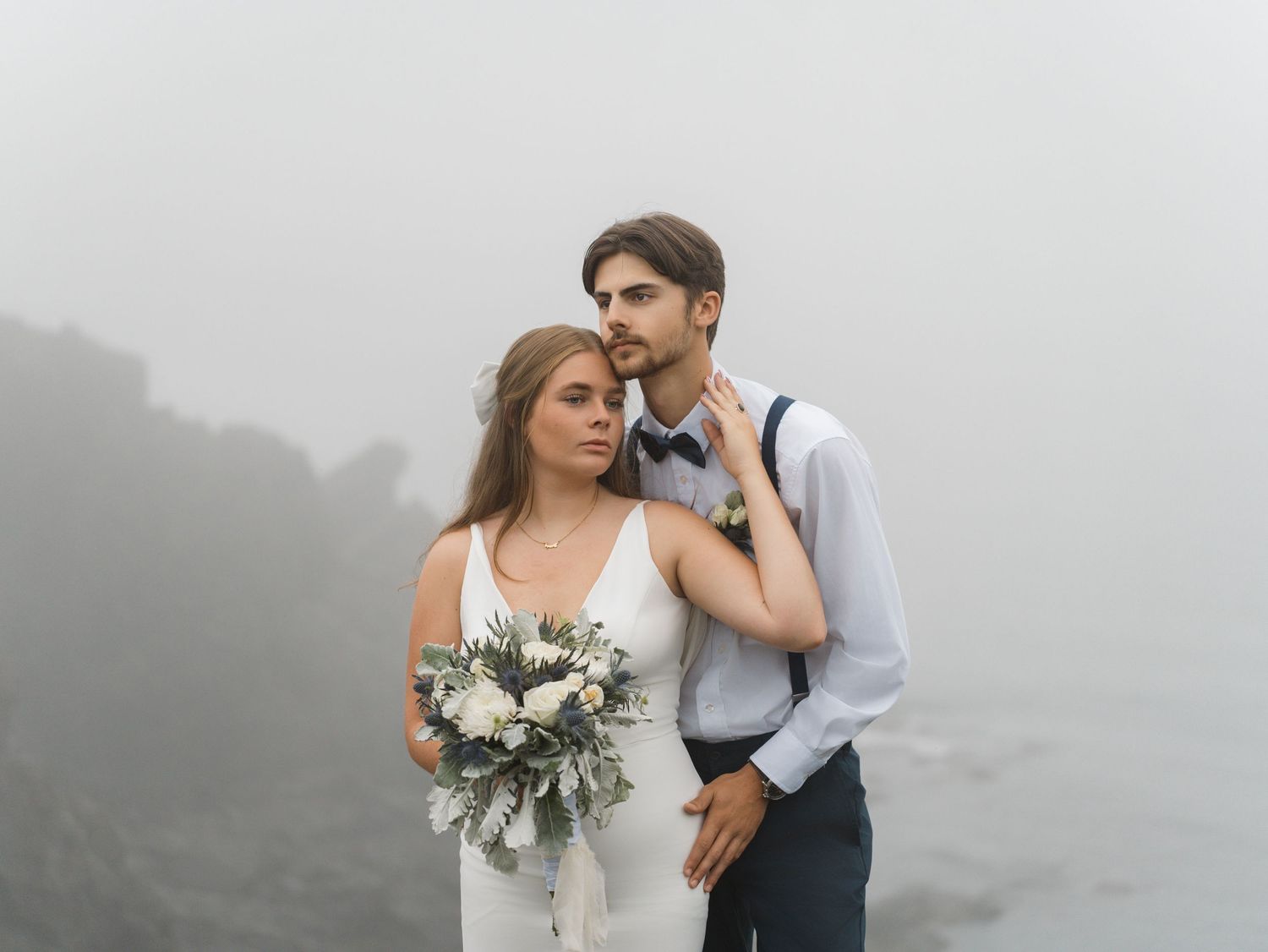 A couple stands together in formal attire on a misty clifftop overlooking the sea.