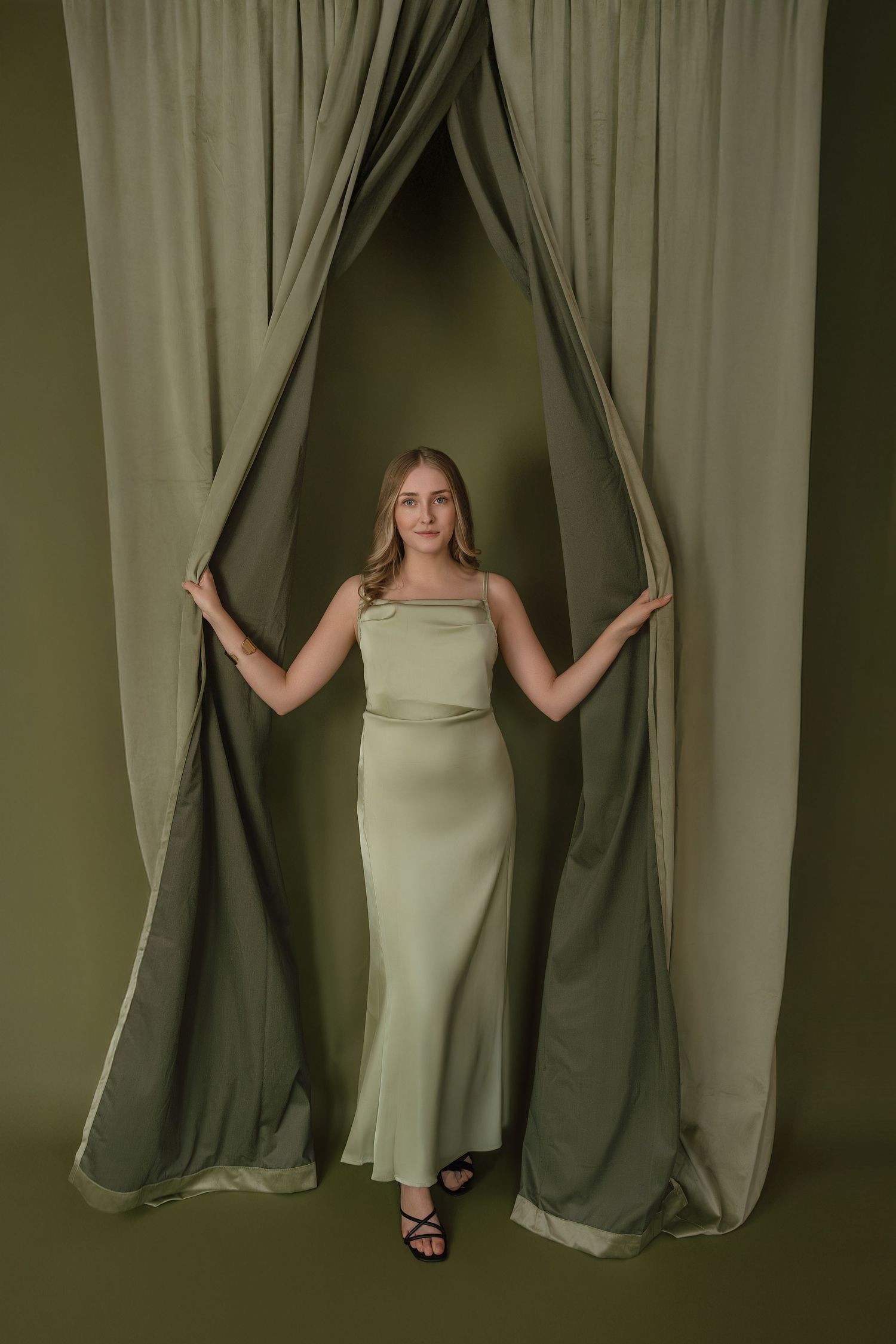 A figure in a light-colored dress emerges through olive green curtains in an artistic fashion portrait series.