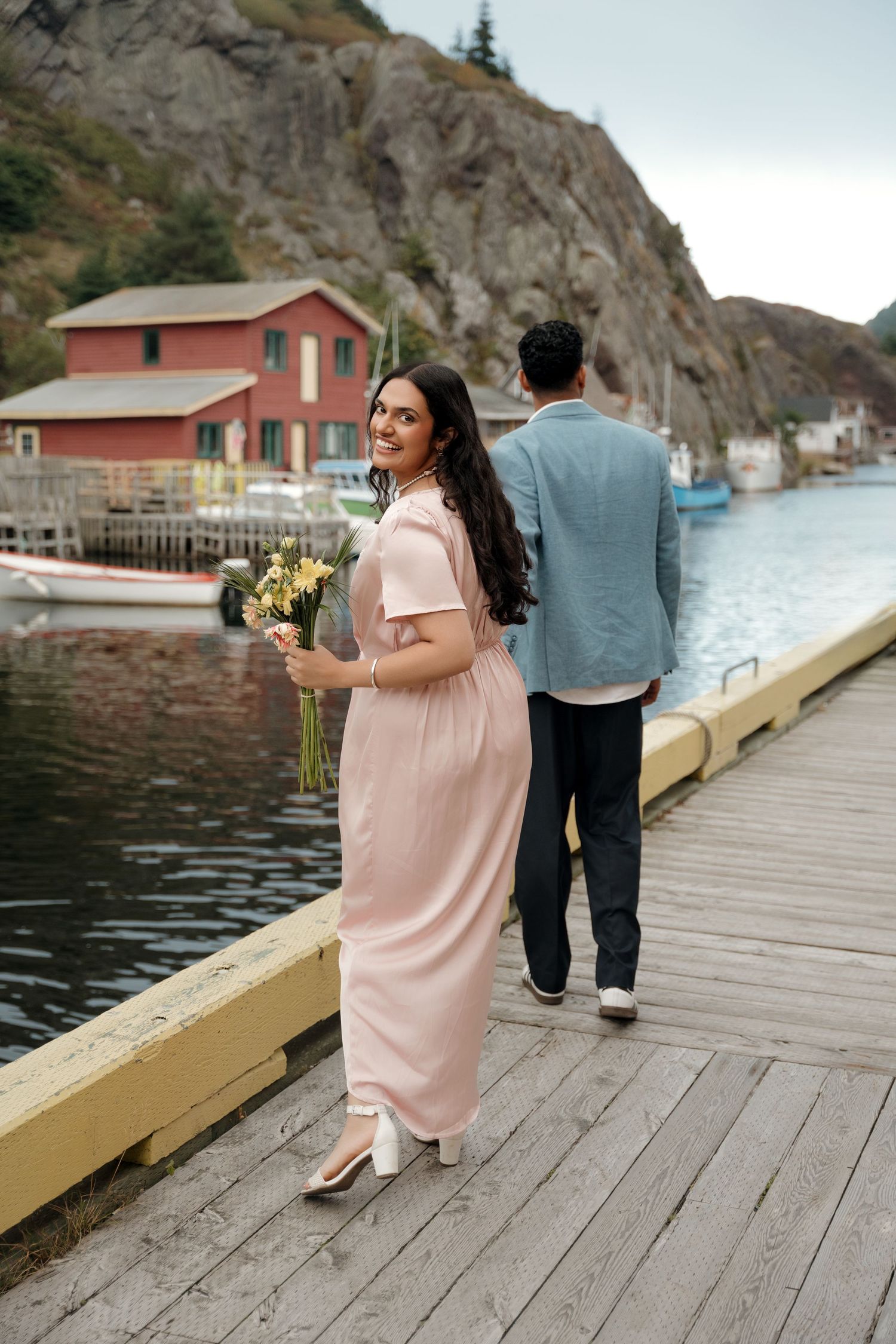 Two people stroll along a waterfront boardwalk near red buildings and rocky cliffs in a scenic coastal setting.