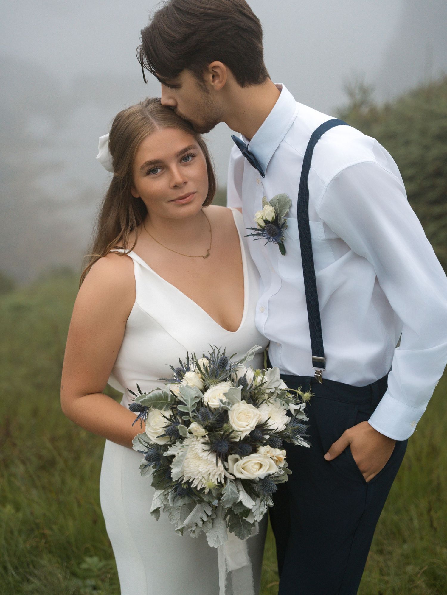 Newlyweds embrace on a hillside with white flowers and navy suspenders against a moody grey backdrop.