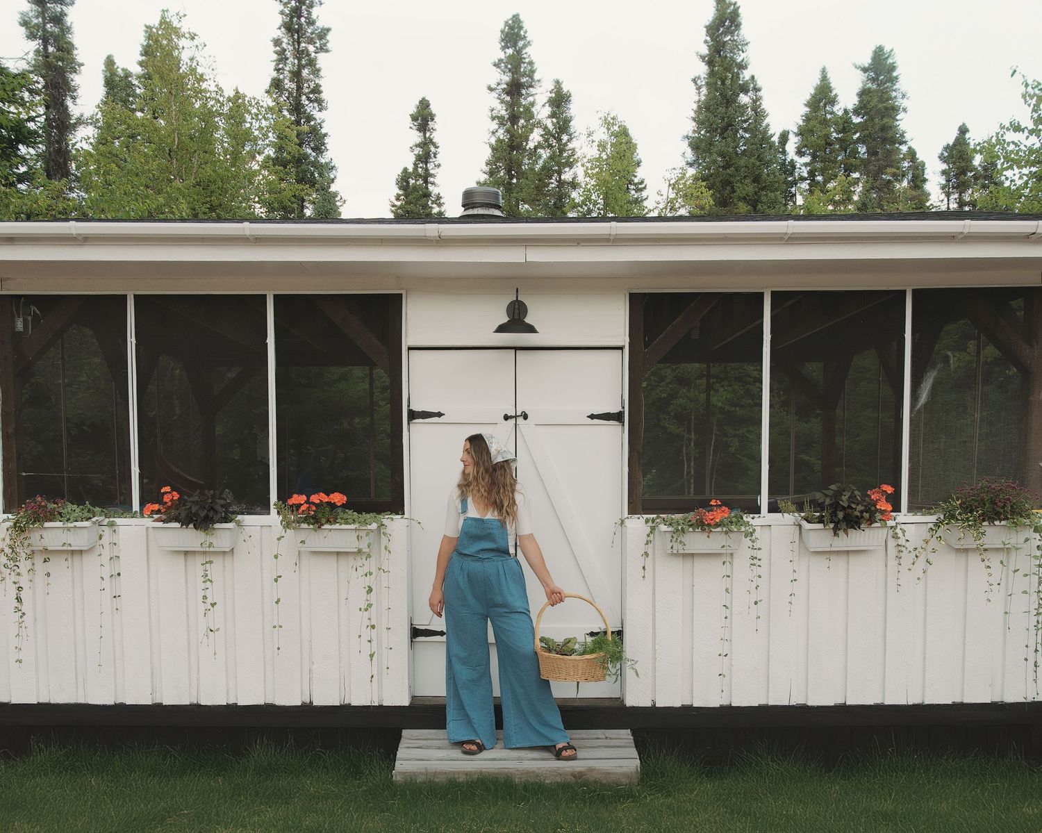 A person in denim overalls standing at the entrance of a white rustic building with hanging plants.