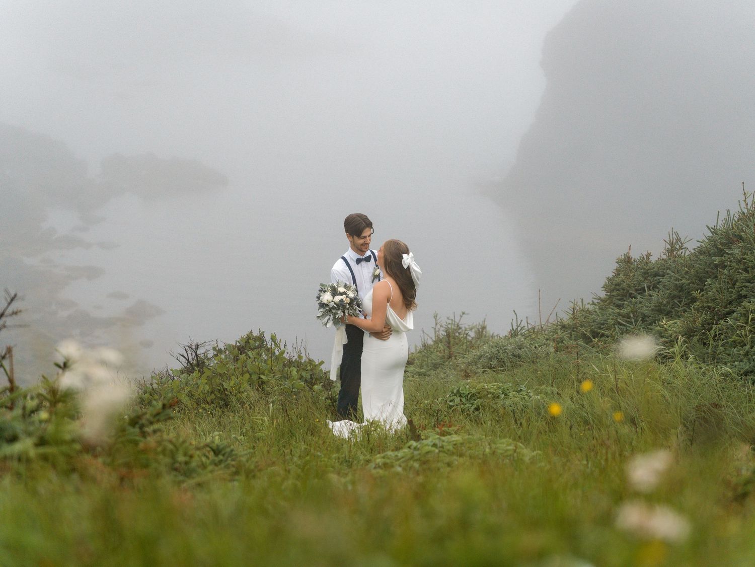 A small wedding couple walks together through a misty meadow surrounded by wildflowers on a foggy summer day.