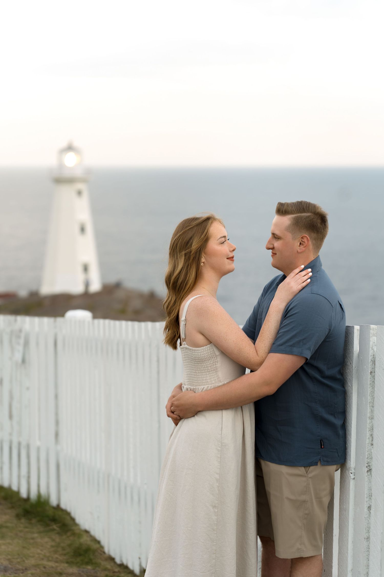 A couple shares tender moments in front of a white picket fence and lighthouse during an outdoor photoshoot.