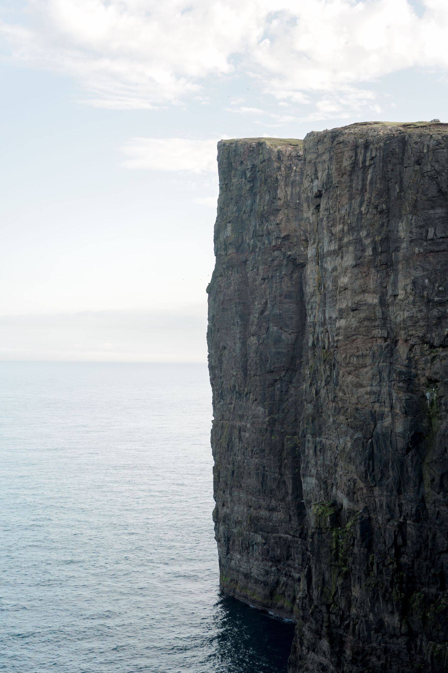 Dramatic vertical sea cliff face rising from dark ocean waters beneath a partly cloudy sky.