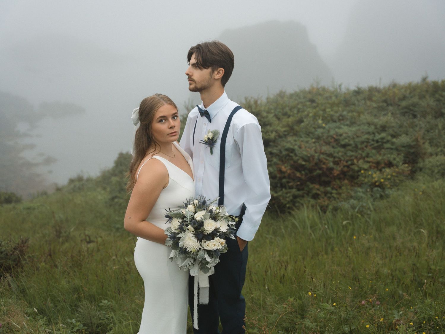 A romantic wedding couple poses together in a misty field with lush greenery and white floral bouquet.