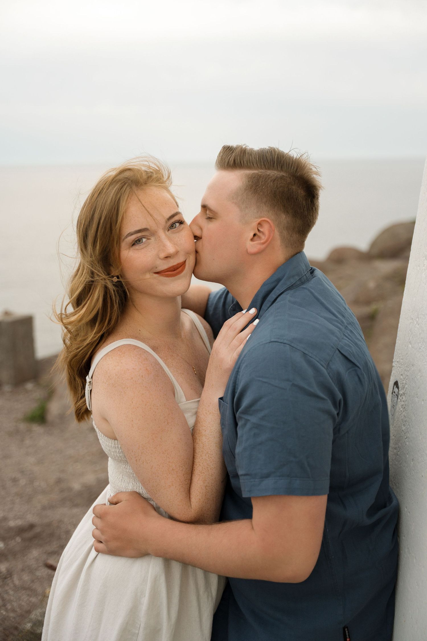 A romantic moment captured between a couple on a cloudy day at the beach with rocky cliffs in the background.