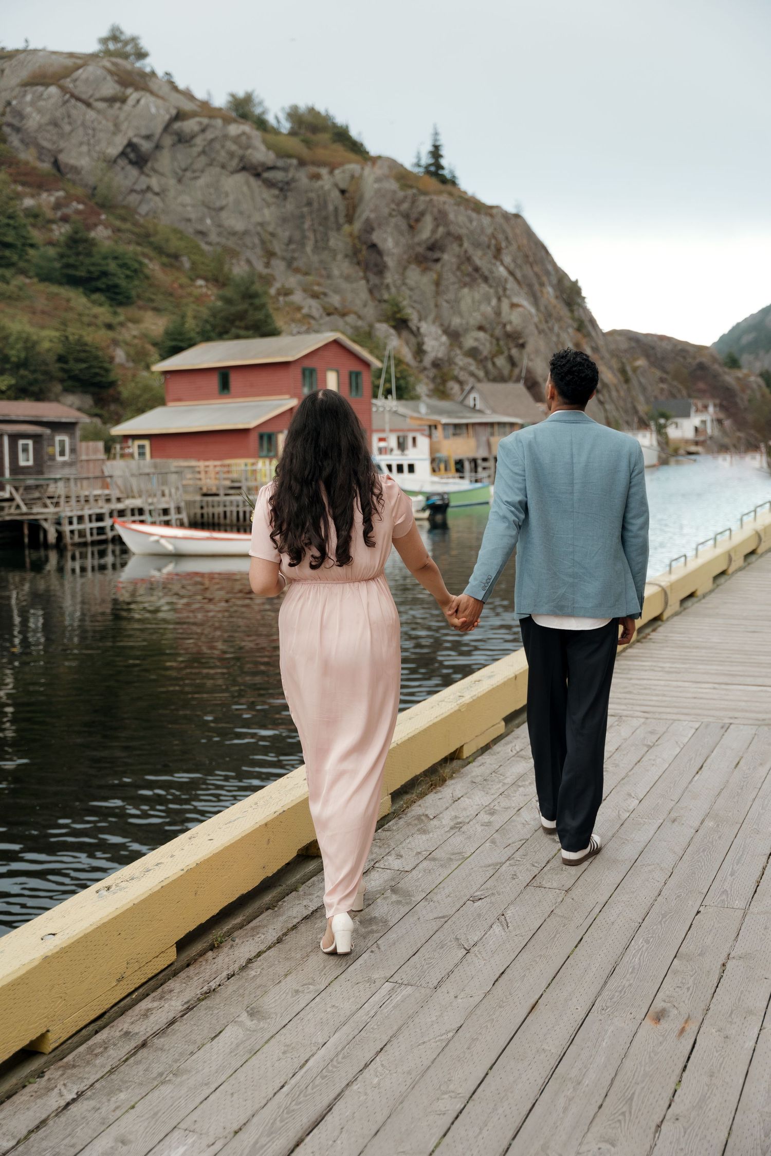 A couple walks along a scenic wooden boardwalk by the water with red buildings and rocky cliffs in the background.