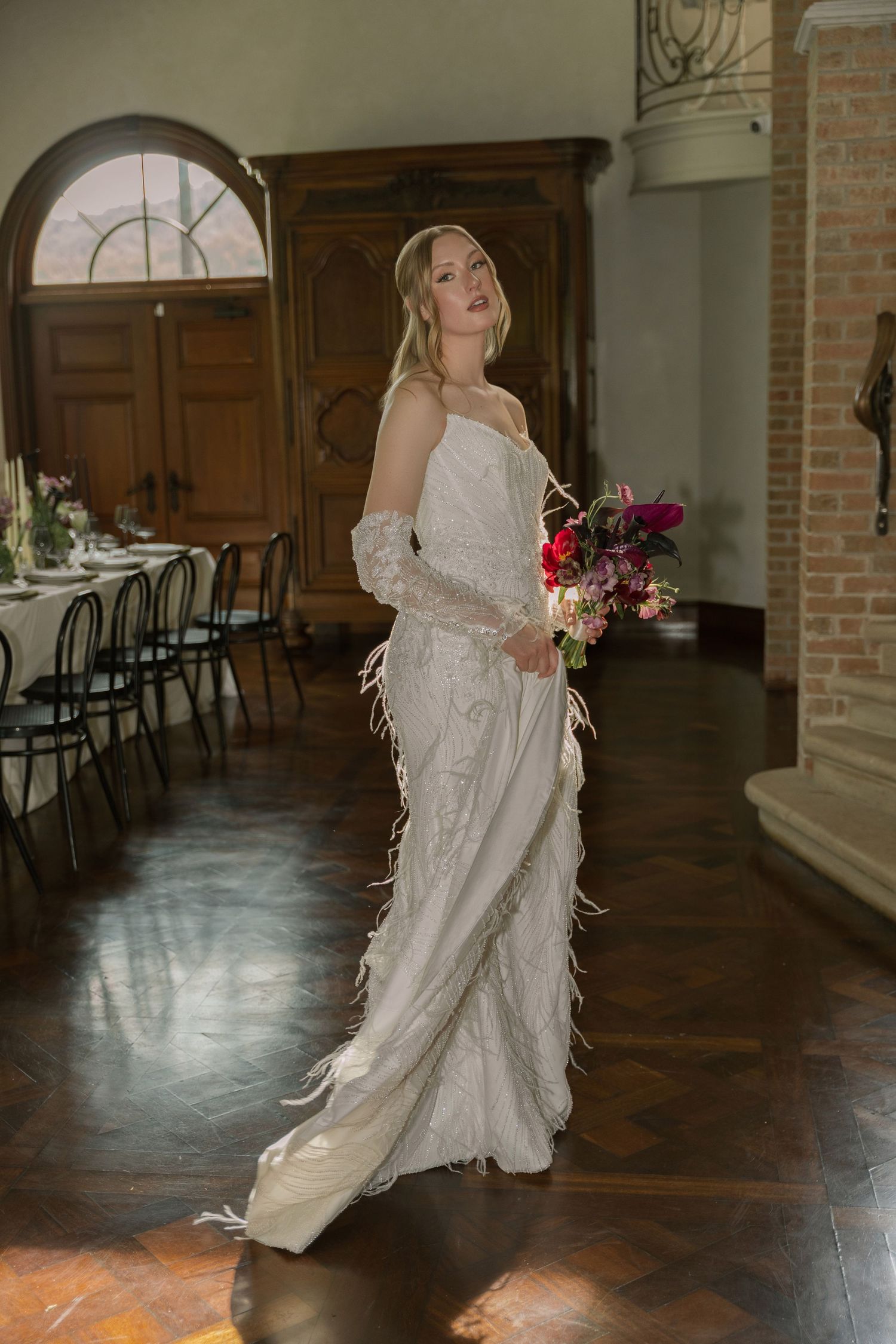 Bride in elegant off-shoulder lace wedding gown with feather details holds vibrant pink bouquet in rustic venue interior.