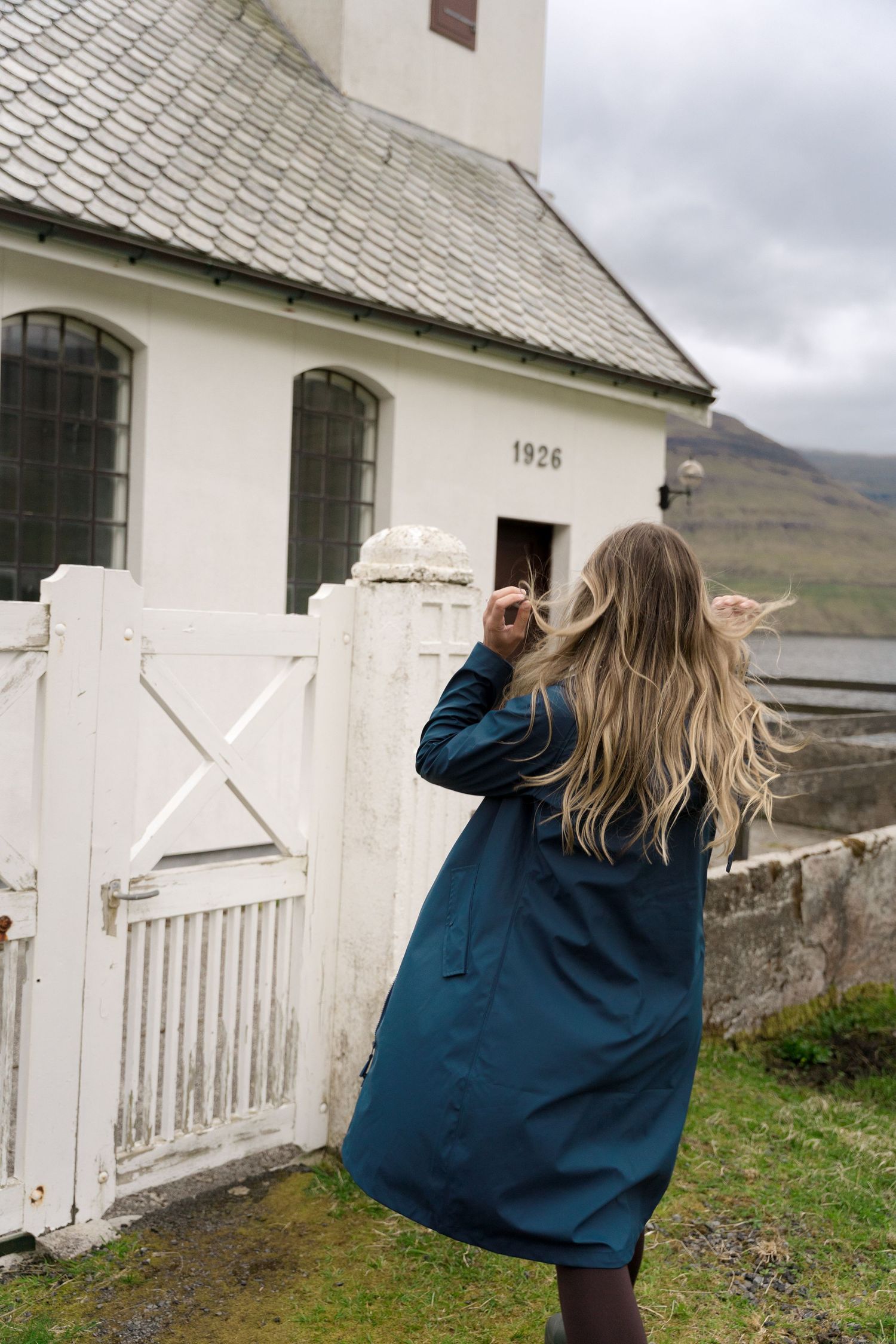 Woman in teal coat with flowing blonde hair stands before a white 1926 church gate on a cloudy day.
