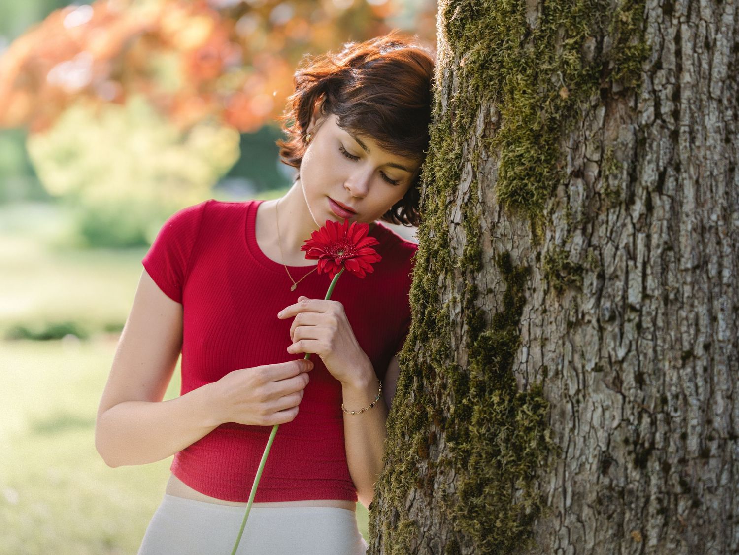 Person in red dress smells a red flower while standing near a tree trunk in a sunny outdoor setting.