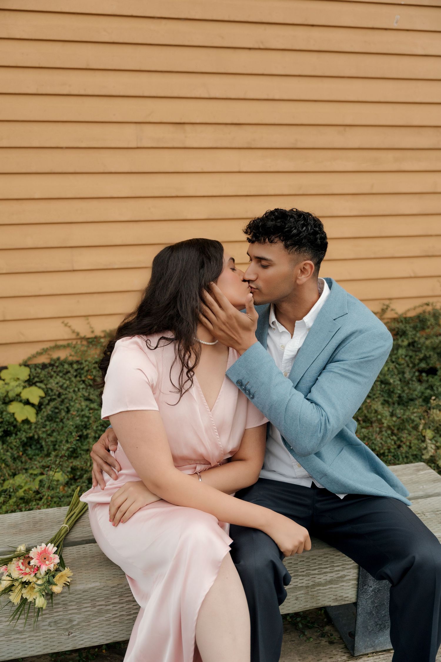 Romantic couple in light pink dress and blue blazer share tender moments on wooden bench during outdoor photo session.