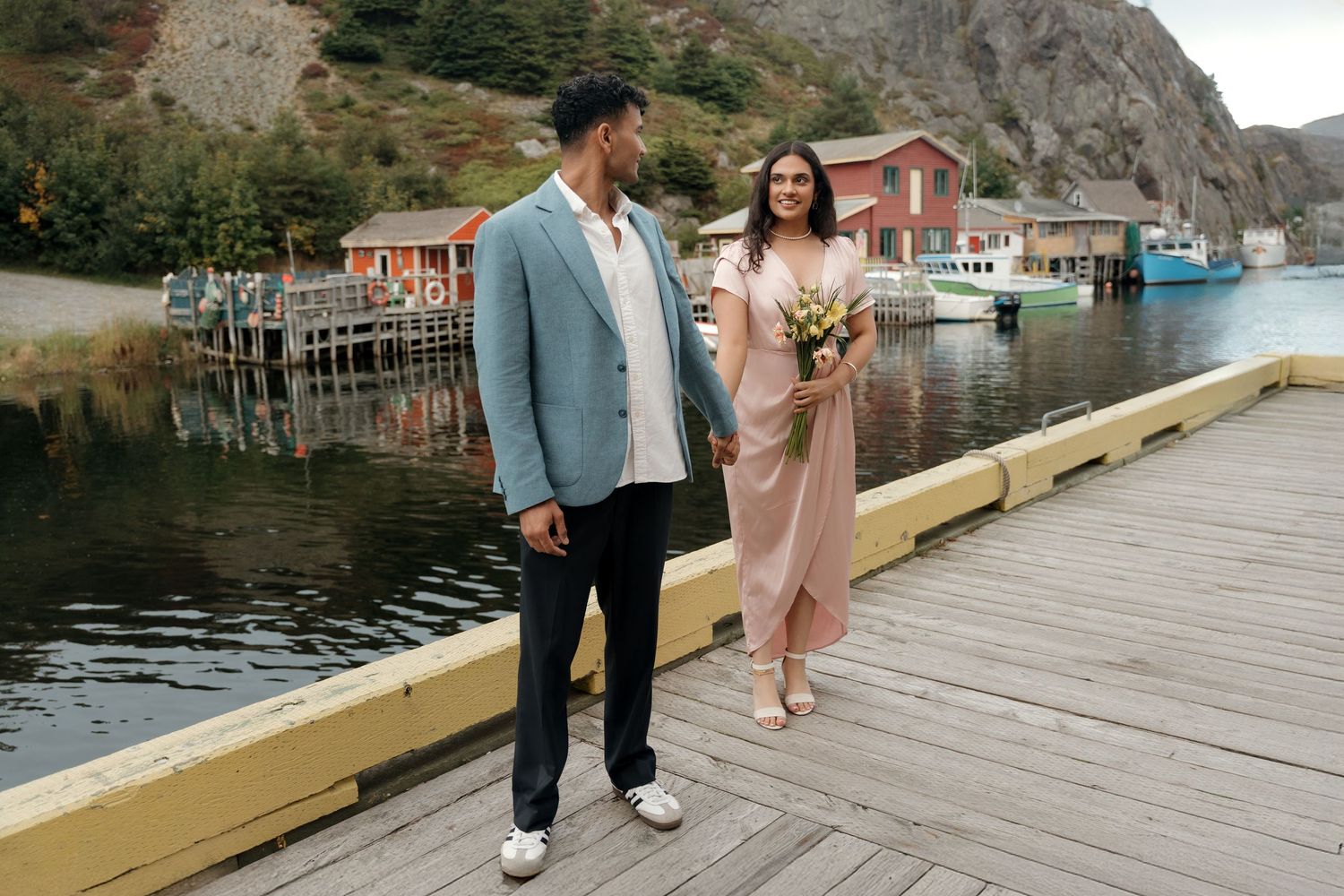 A couple stands on a wooden dock in front of colorful fishing buildings and boats in Newfoundland.