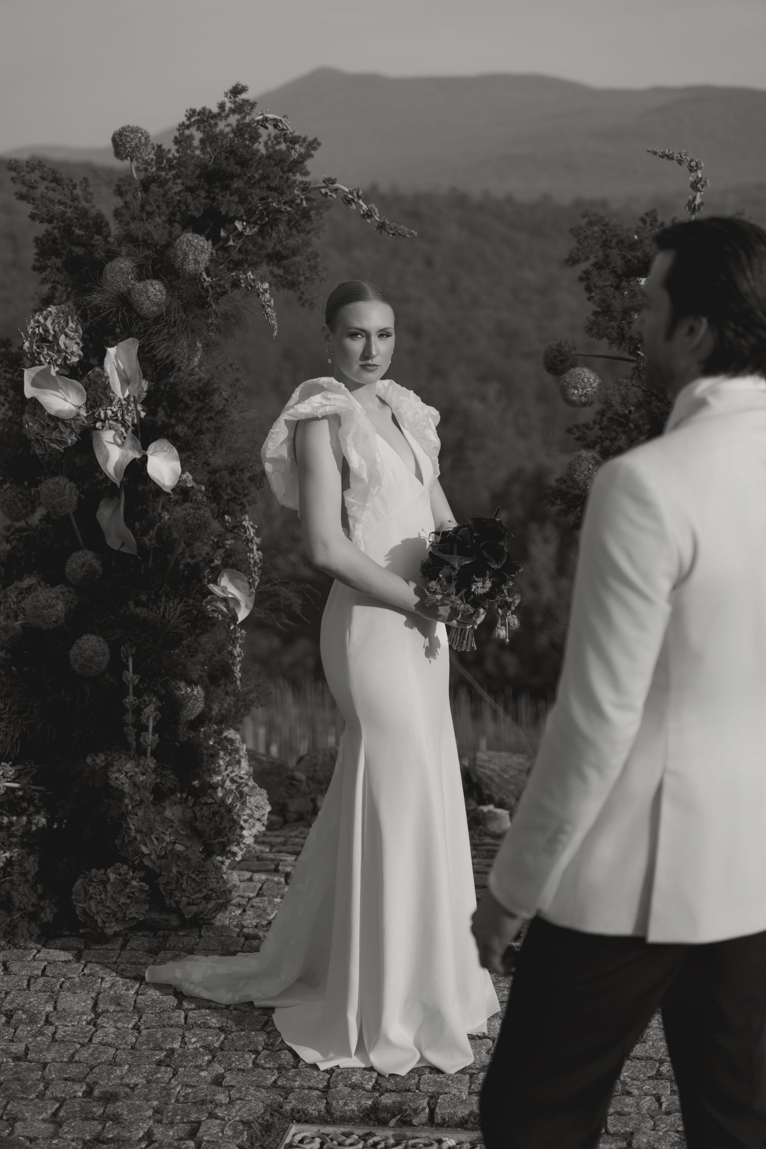 A couple in formal attire embrace during what appears to be an outdoor wedding ceremony with mountains in the background.