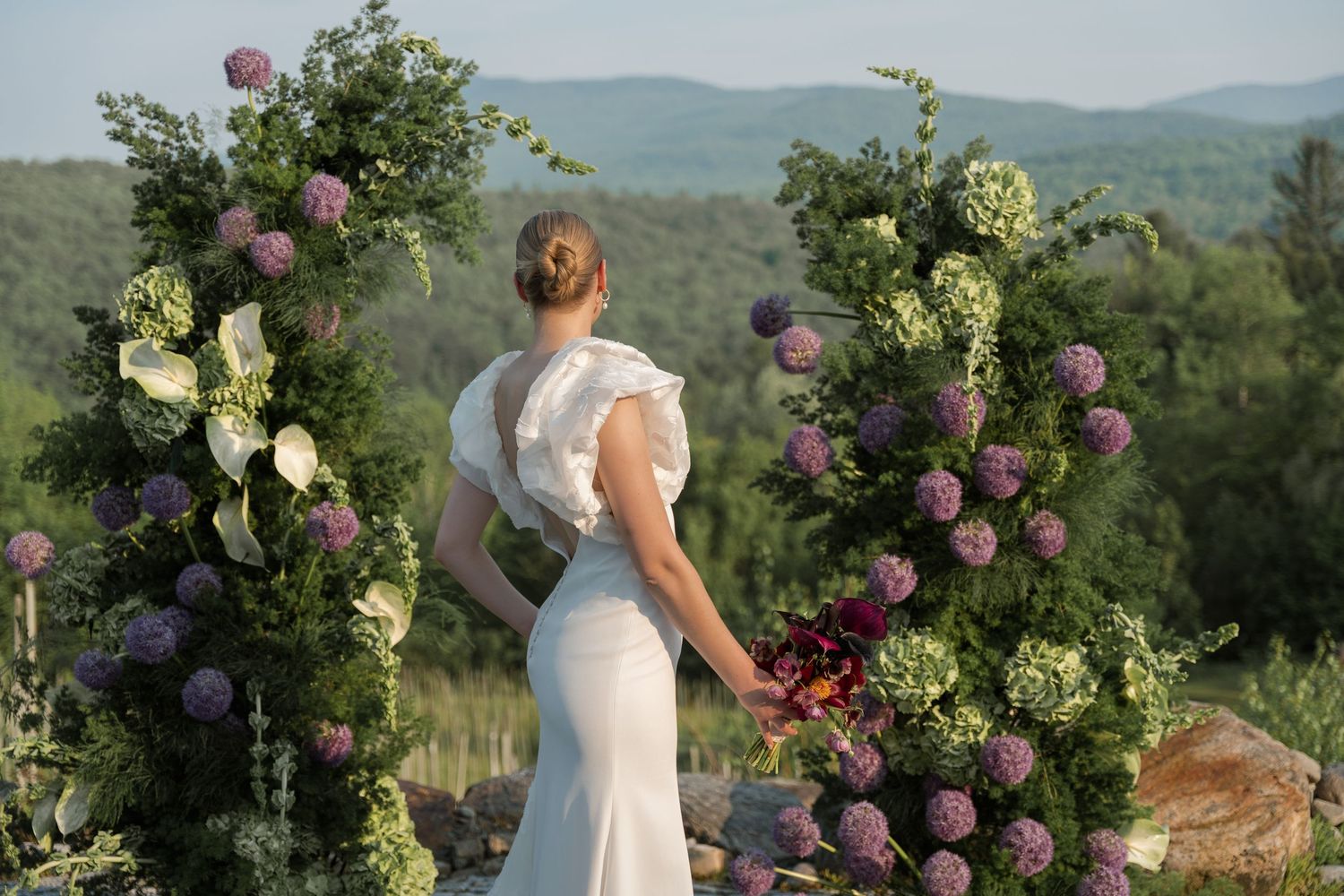 Summer wedding ceremony on a hilltop adorned with blooming pink and white floral arrangements and mountains in background.